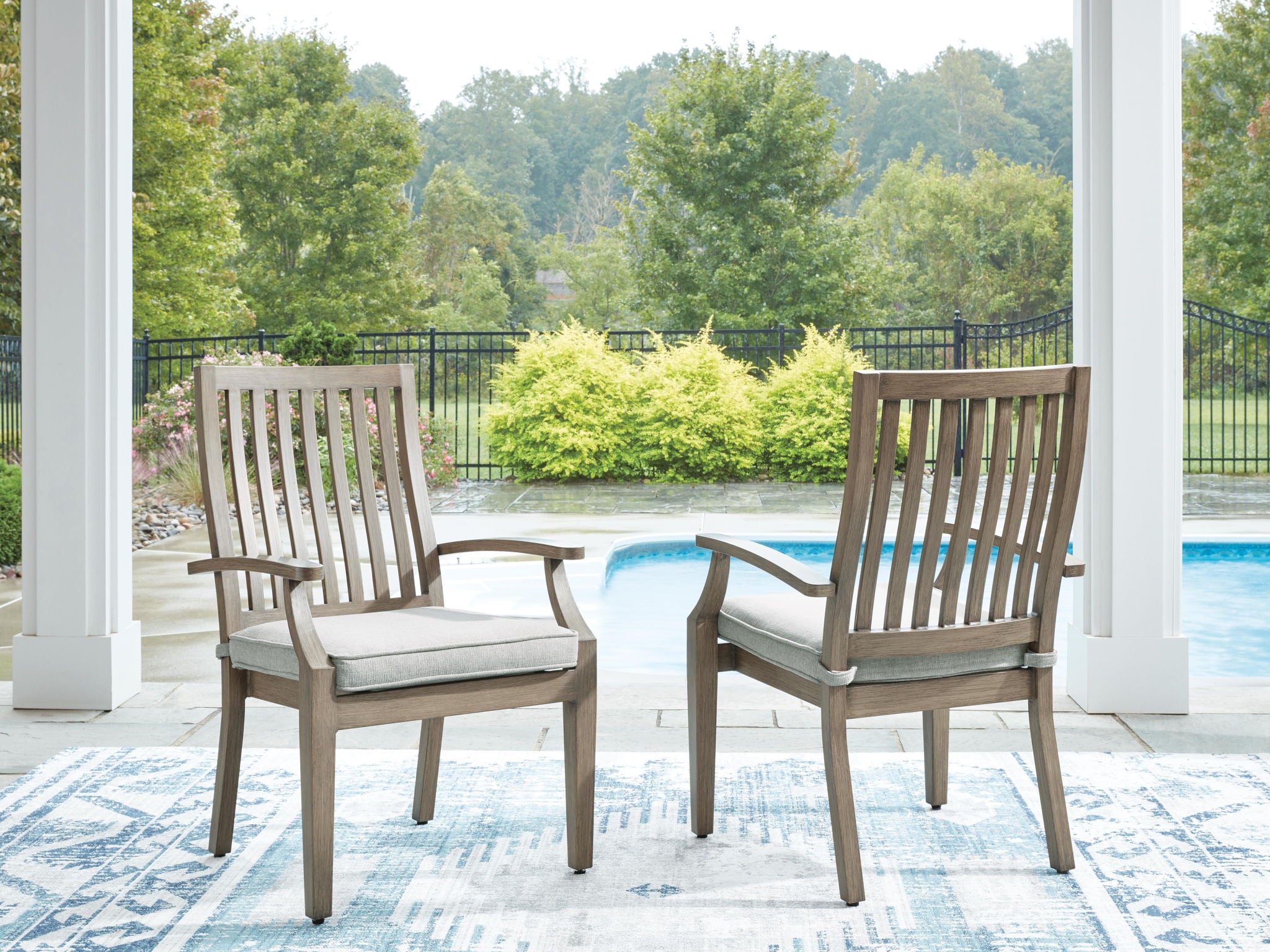 Two wooden arm chairs with slatted backs and beige seat cushions on patio.