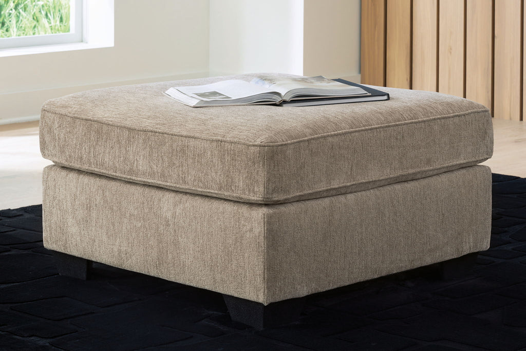 Light brown ottoman with cushioned top and dark legs, shown with a folded magazine