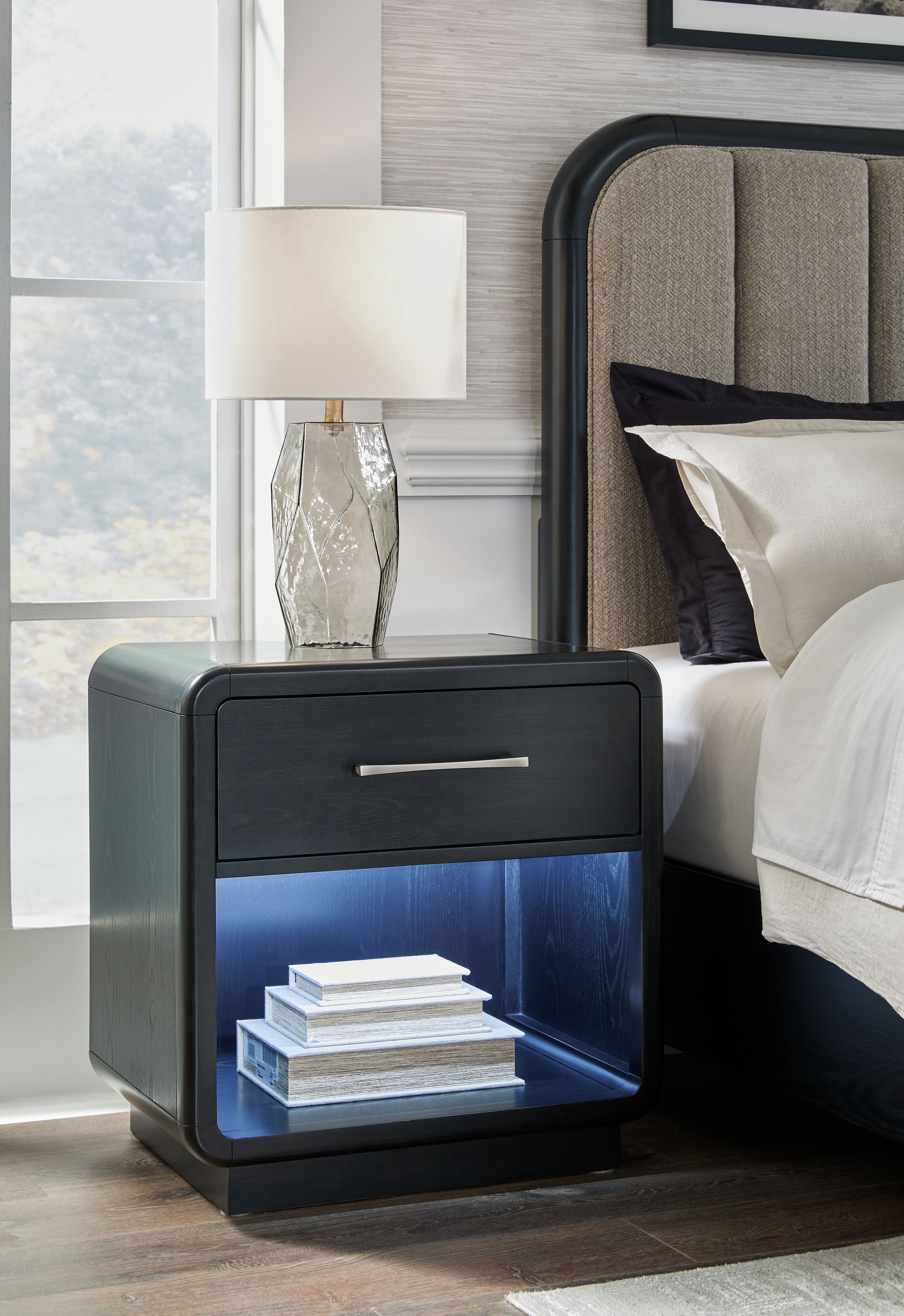 Black night stand with one drawer and open shelf holding stacked books beside bed
