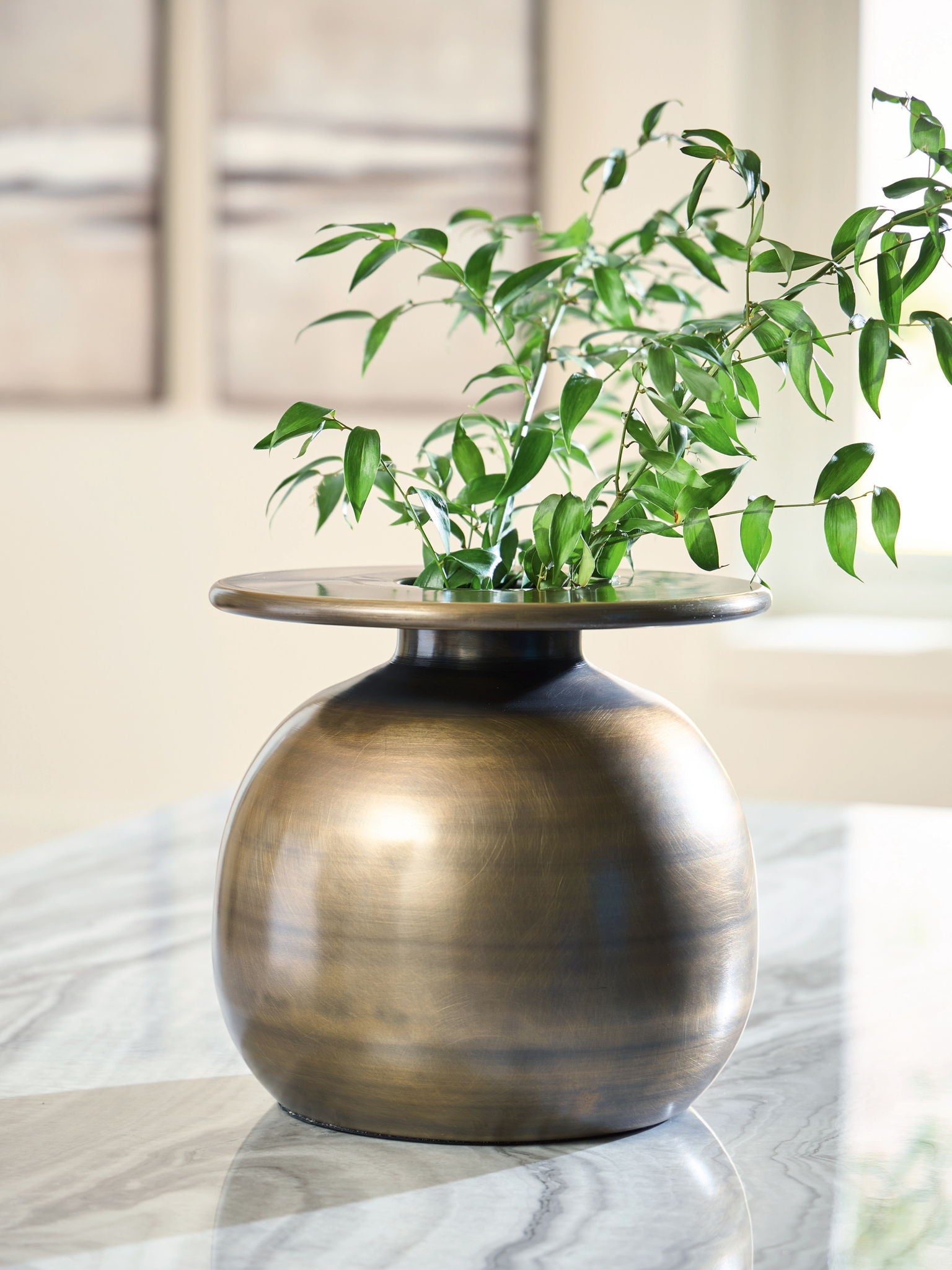 Bronze metal vase centerpiece with wide mouth on round stand, holding green stems and leaves.