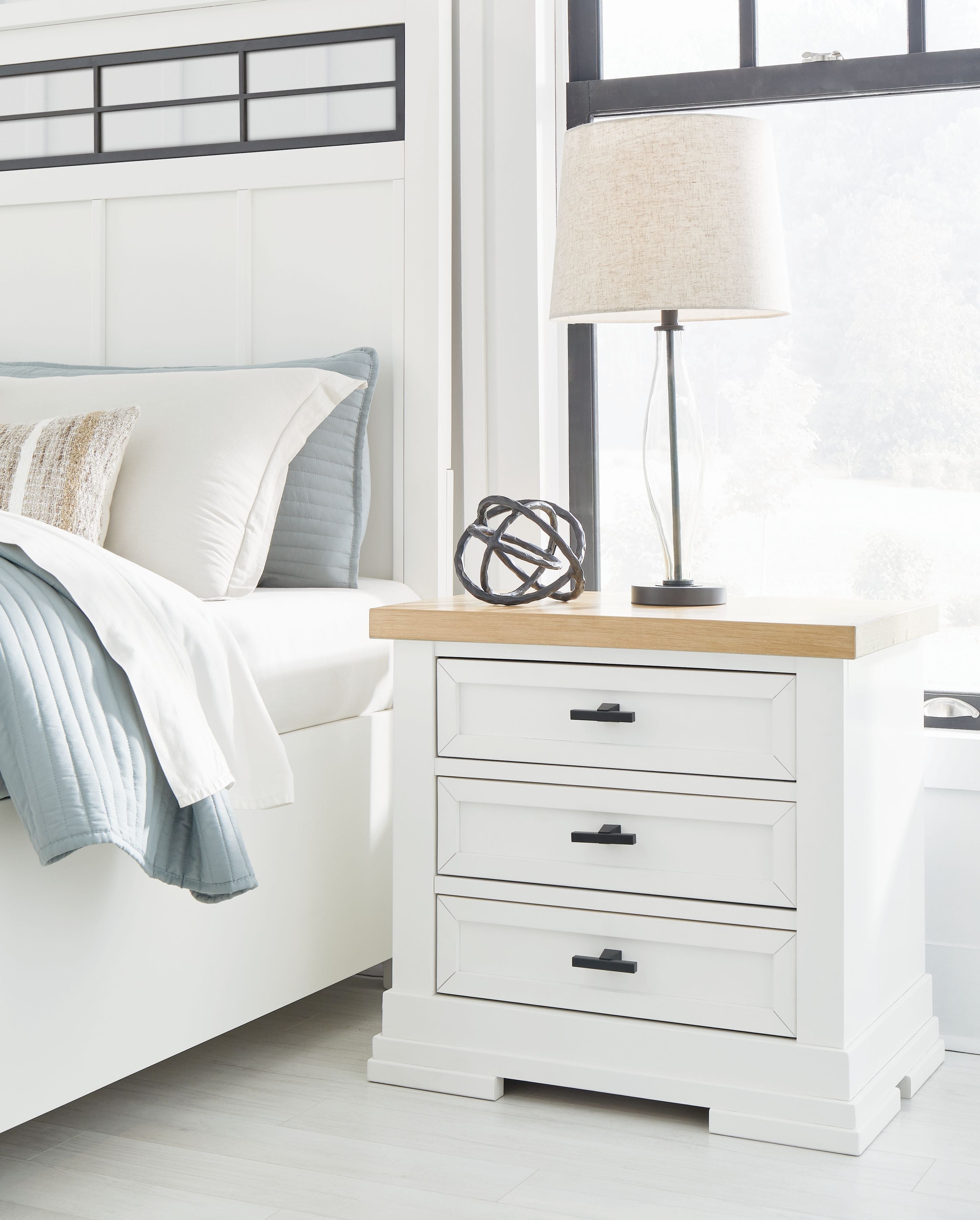 White three-drawer night stand with natural wood top and black handles beside lamp