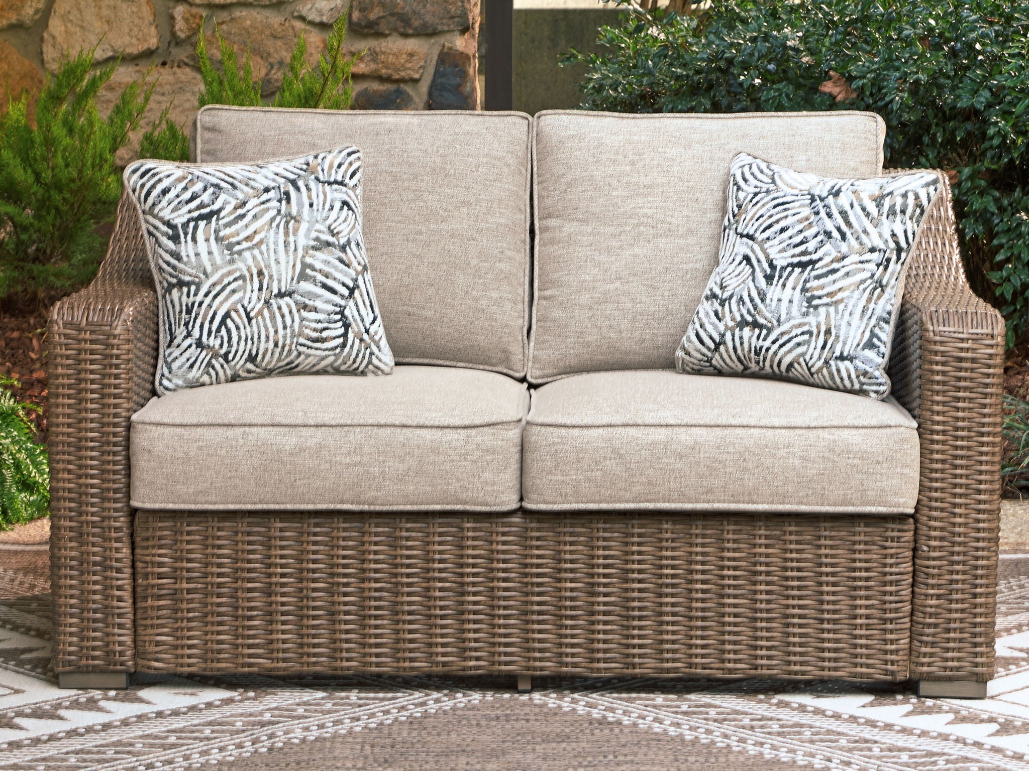Outdoor loveseat with beige seat cushions, brown wicker frame, two patterned throw pillows
