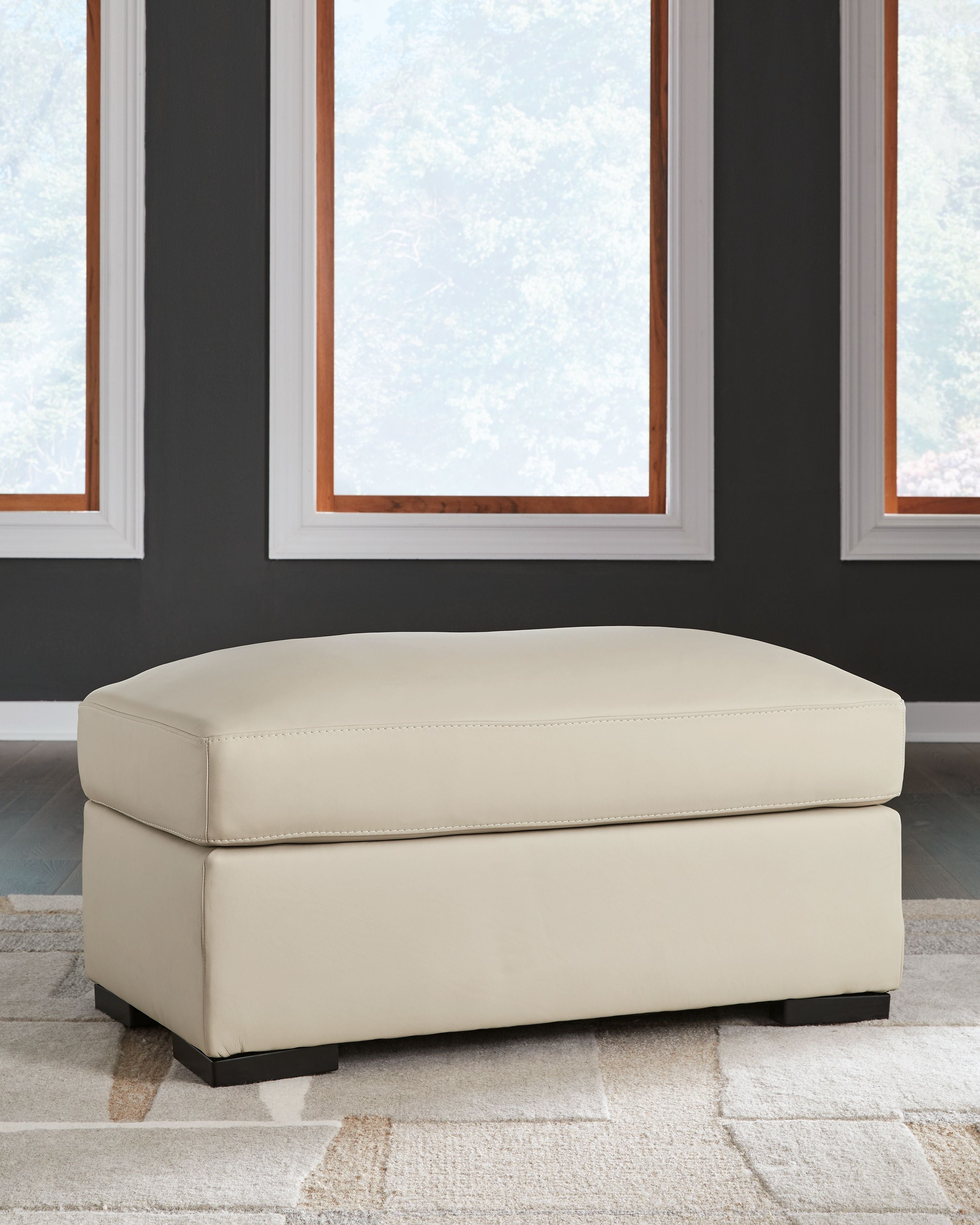 Almond ottoman with padded lid and sides, stitched trim, and dark rectangular feet.