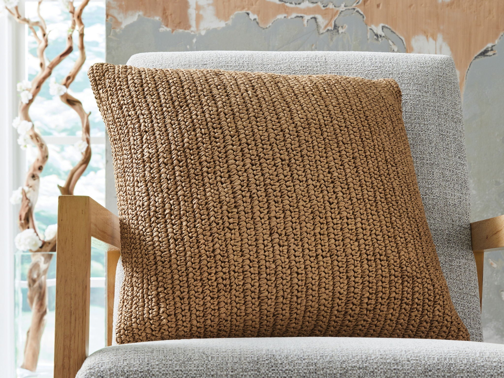 Tan woven decorative pillow on gray chair, square shape with stitched edge