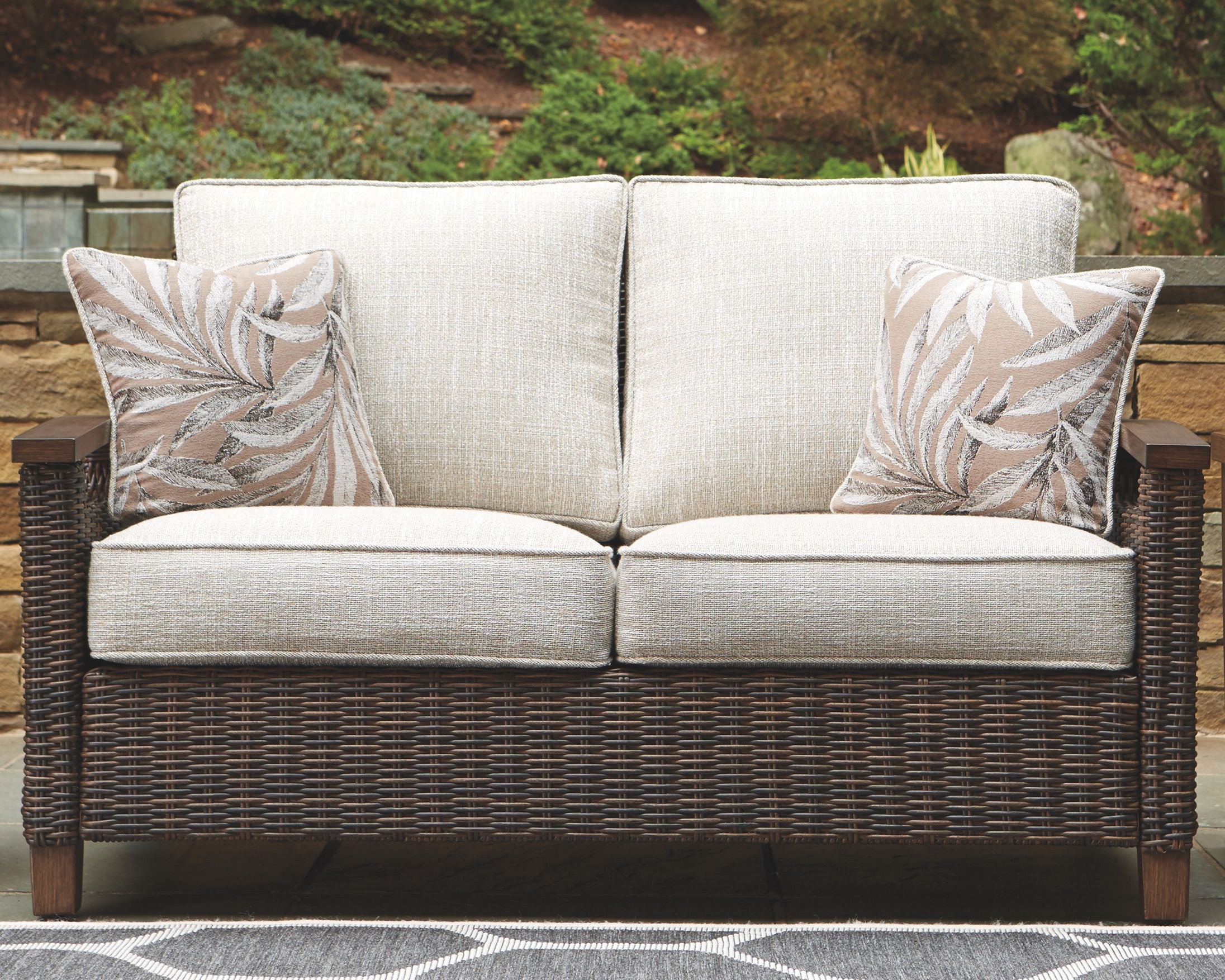 Outdoor loveseat with cushion, light beige upholstery, dark wicker base, leaf-pattern pillows