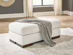 Cream upholstered ottoman with dark wooden legs, gray throw blanket draped on top