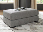 Grey fabric ottoman with thick square seat and black legs, styled with magazine on top