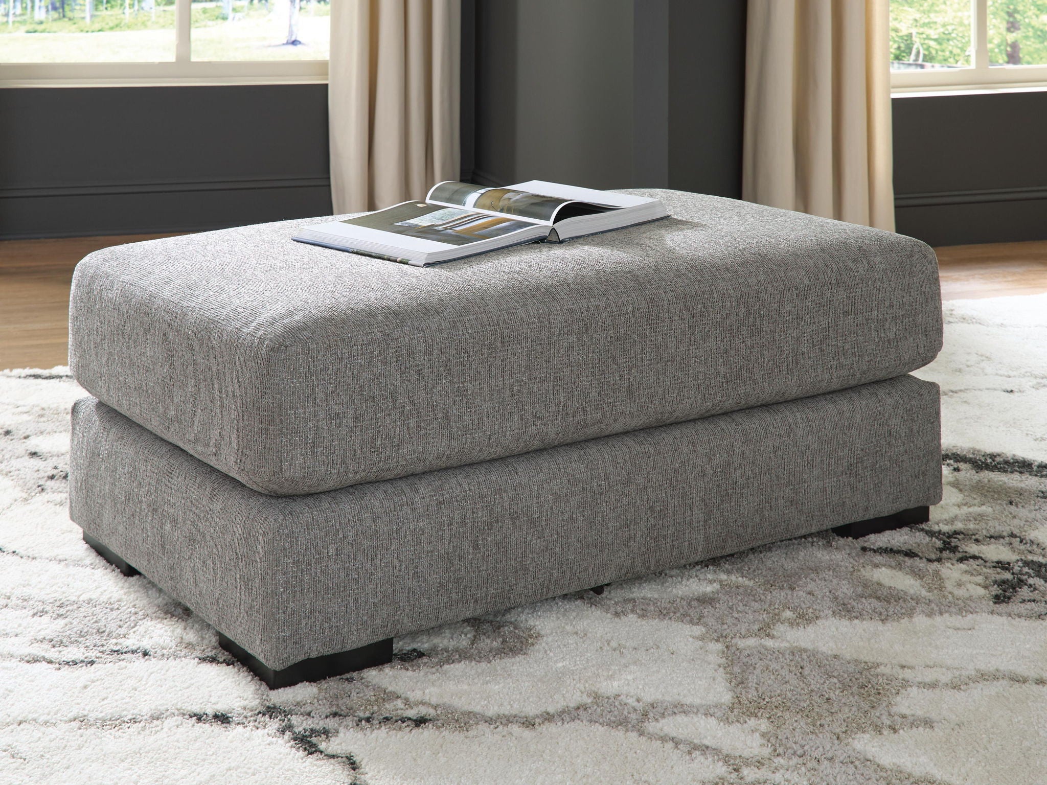 Grey fabric ottoman with thick square seat and black legs, styled with magazine on top