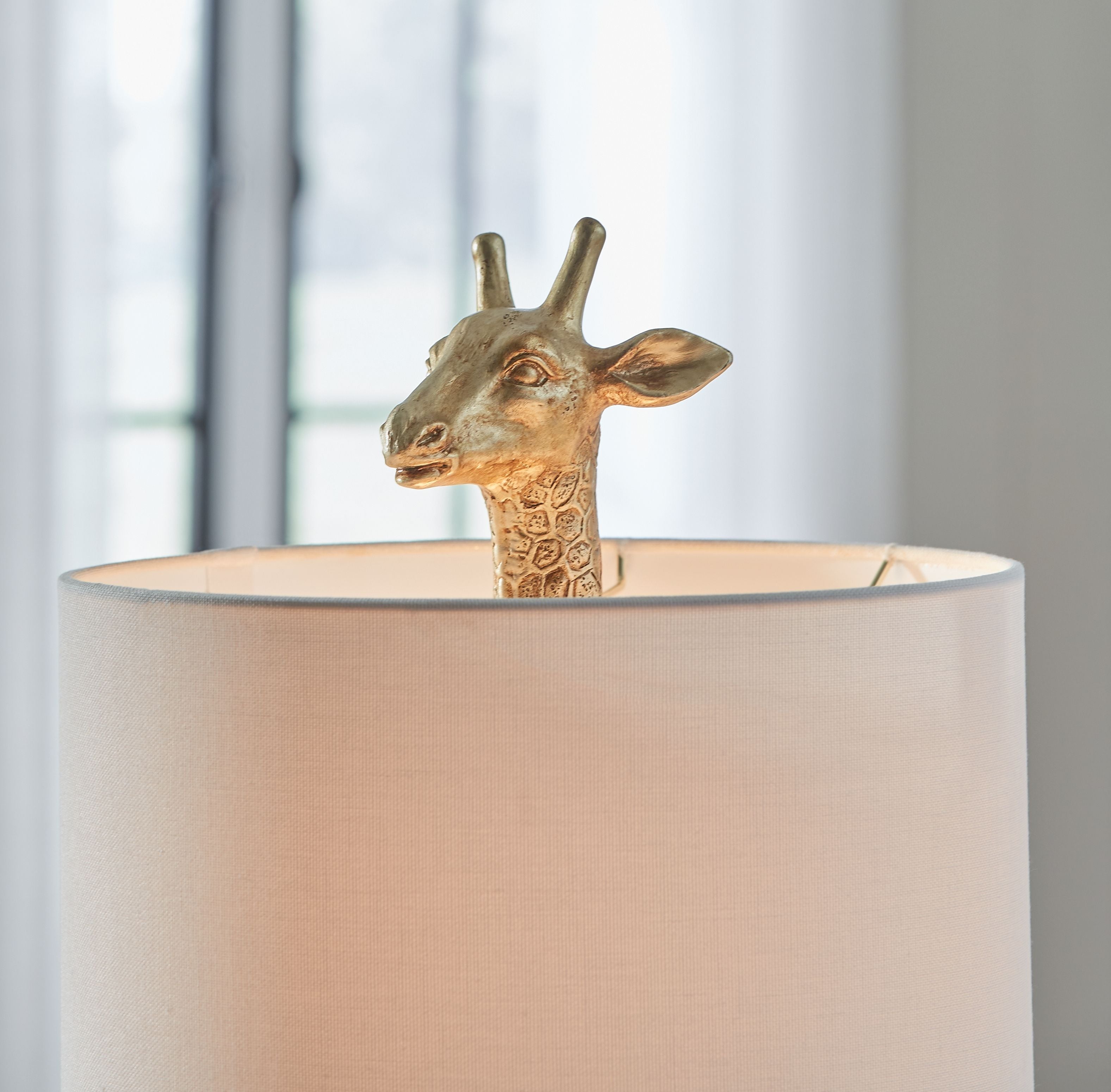 Gold table lamp with giraffe head detail and white fabric drum shade.