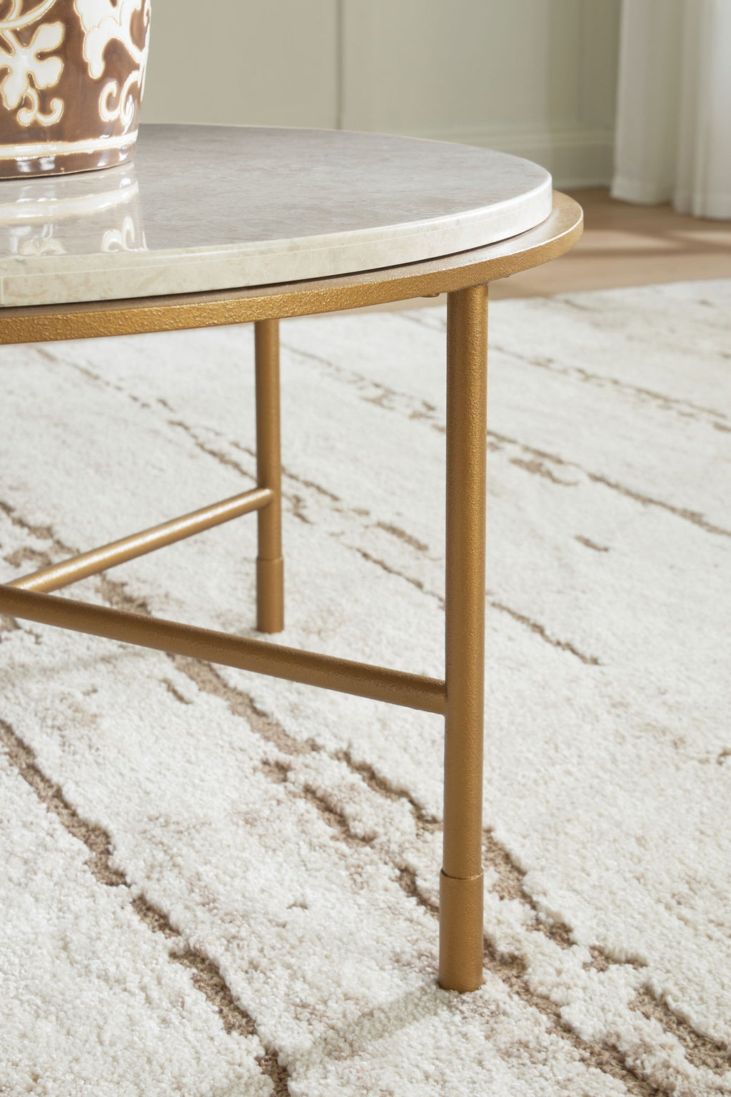 Round table corner view with cream stone-look top and gold metal cross-brace legs.