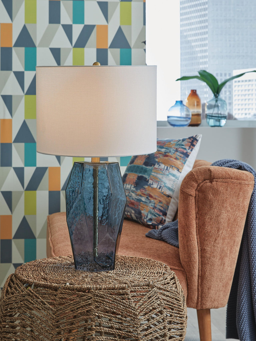 Blue glass table lamp with white drum shade and faceted body, standing on rug