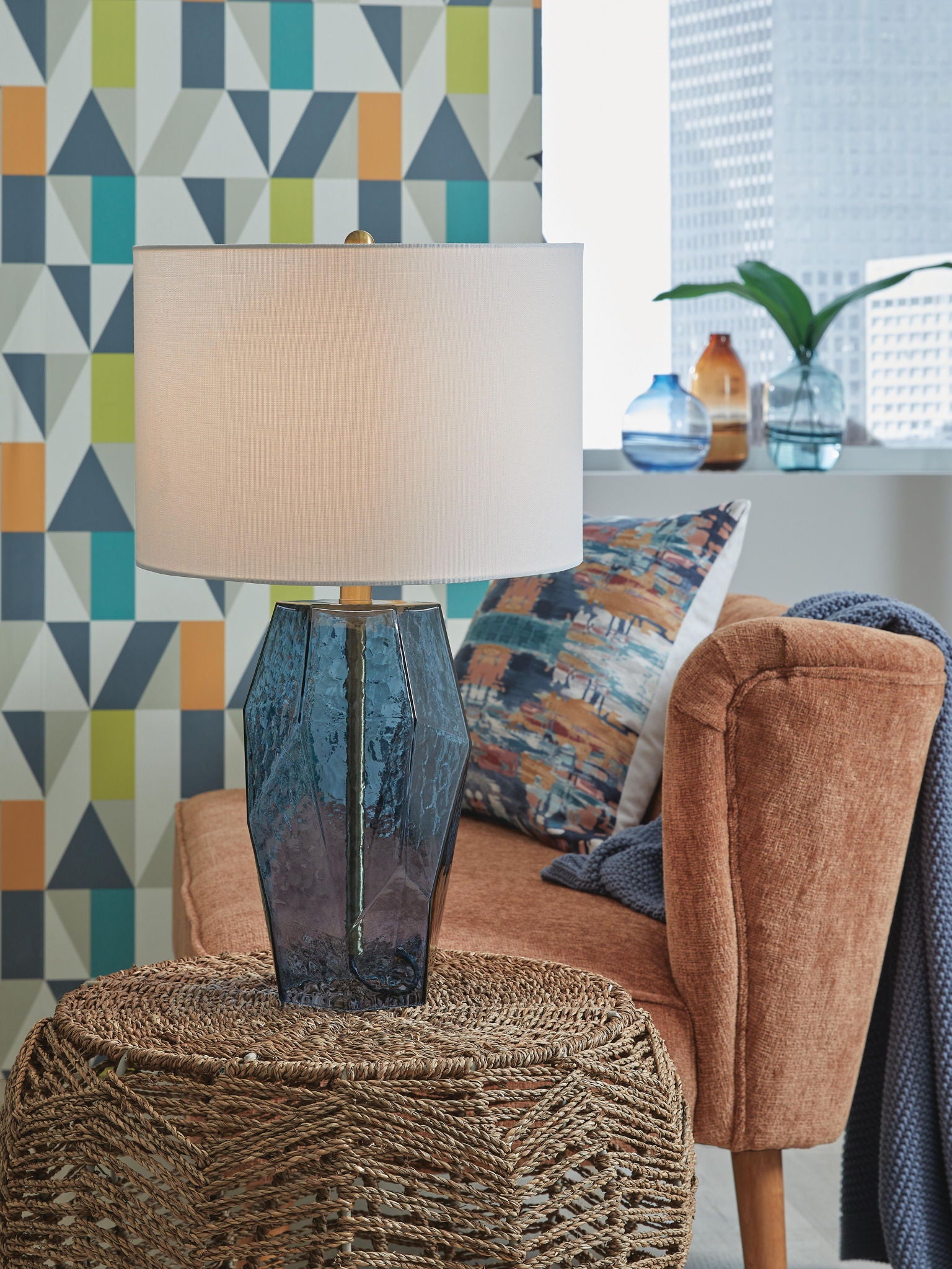 Blue glass table lamp with white drum shade and faceted body, standing on rug