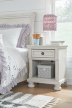 Antique white night stand with one drawer and open shelf next to a bed