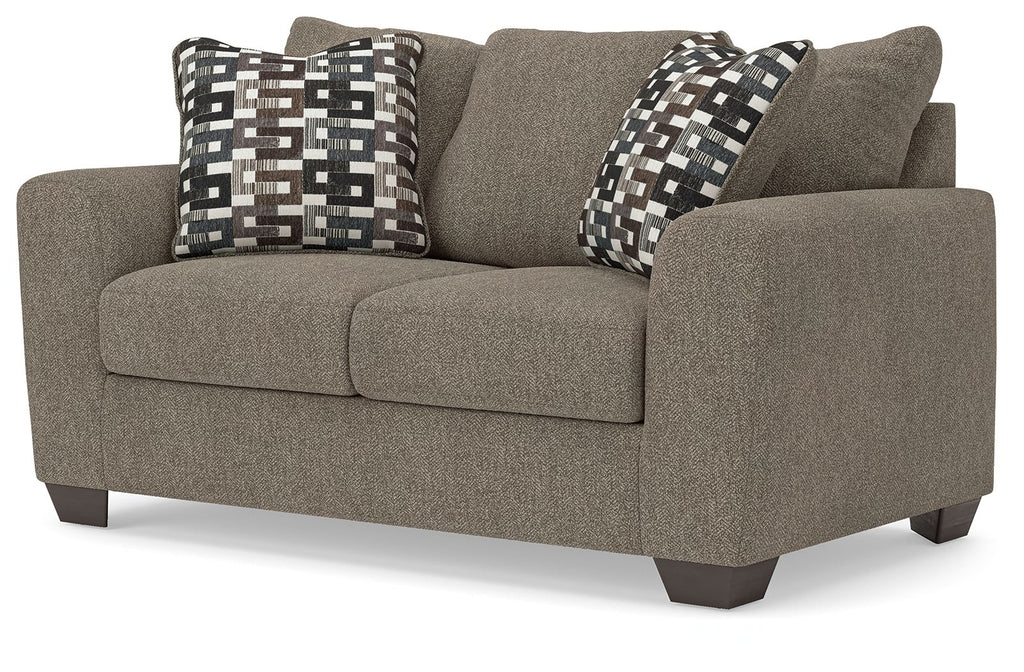 Grey suede loveseat with padded arms and two patterned throw pillows