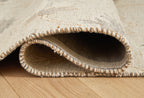 Beige rug edge detail with curved overlap and fringed trim, on wood floor