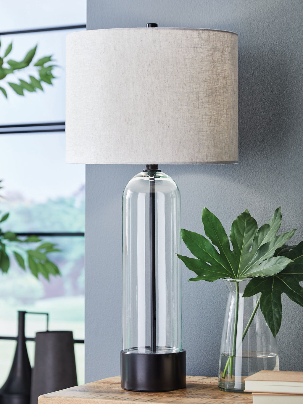 Clear glass table lamp with black base, linen drum shade, and metal stem