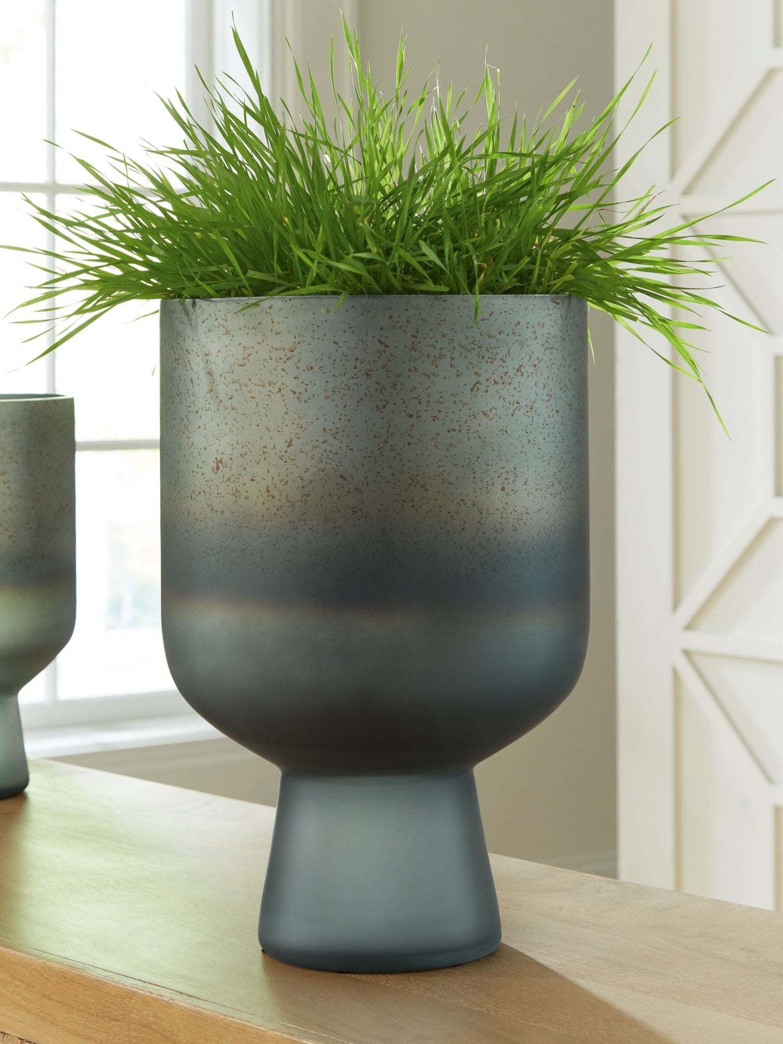 Blue speckled vase holding green grass, flared opening on pedestal base.