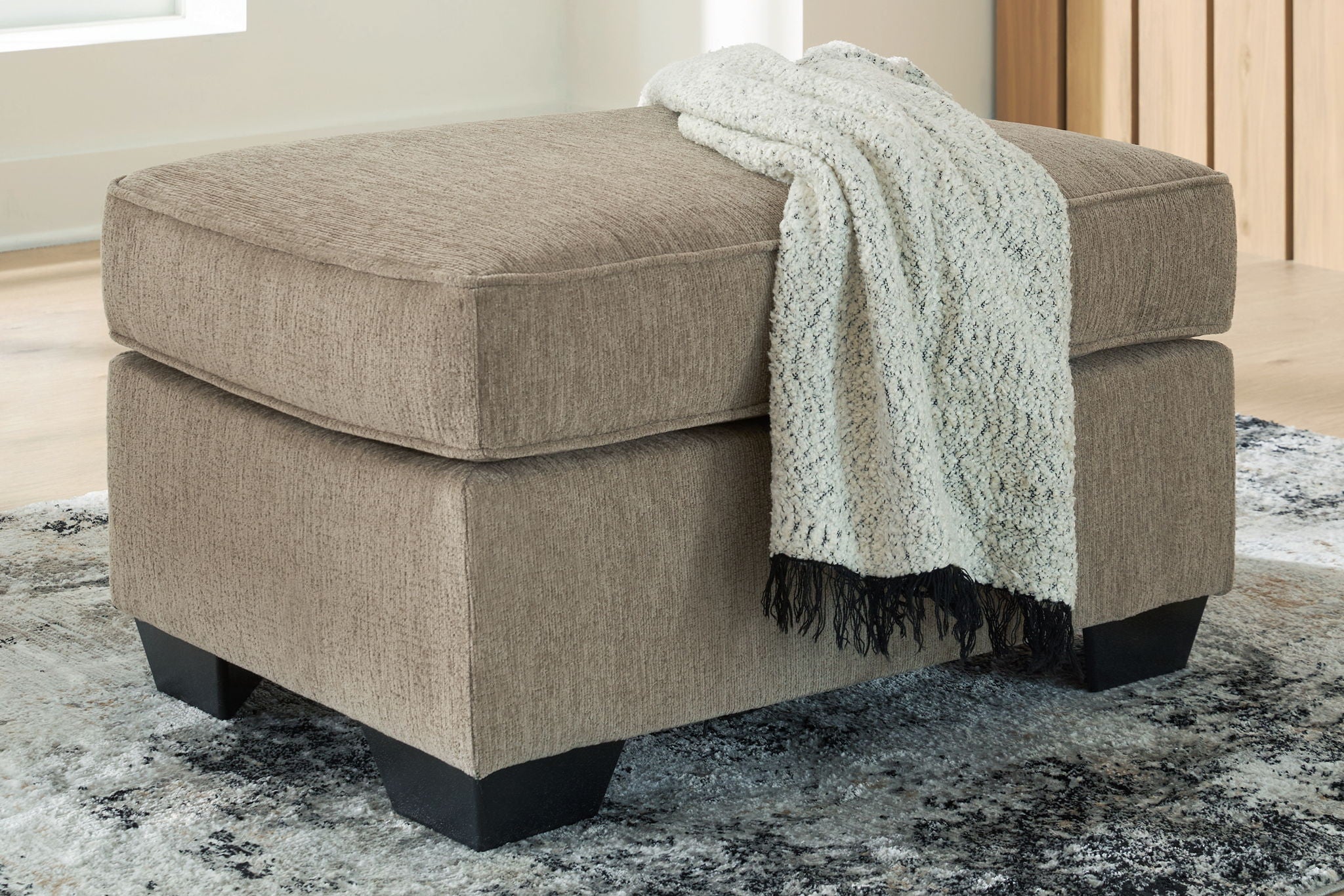 Light brown ottoman with dark legs, styled with a light throw blanket
