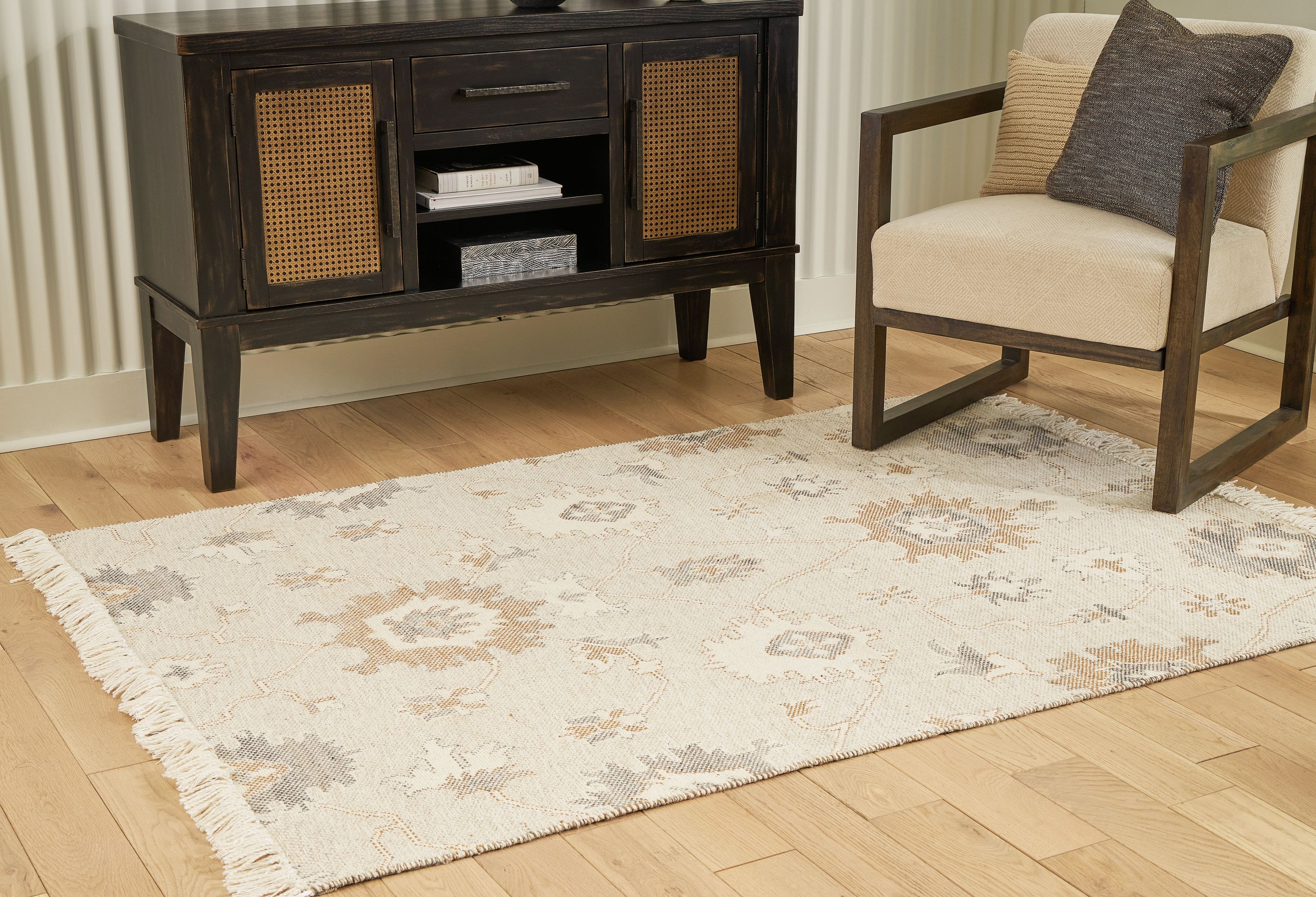 Beige rug with ornate pattern and fringed edges under a chair and cabinet