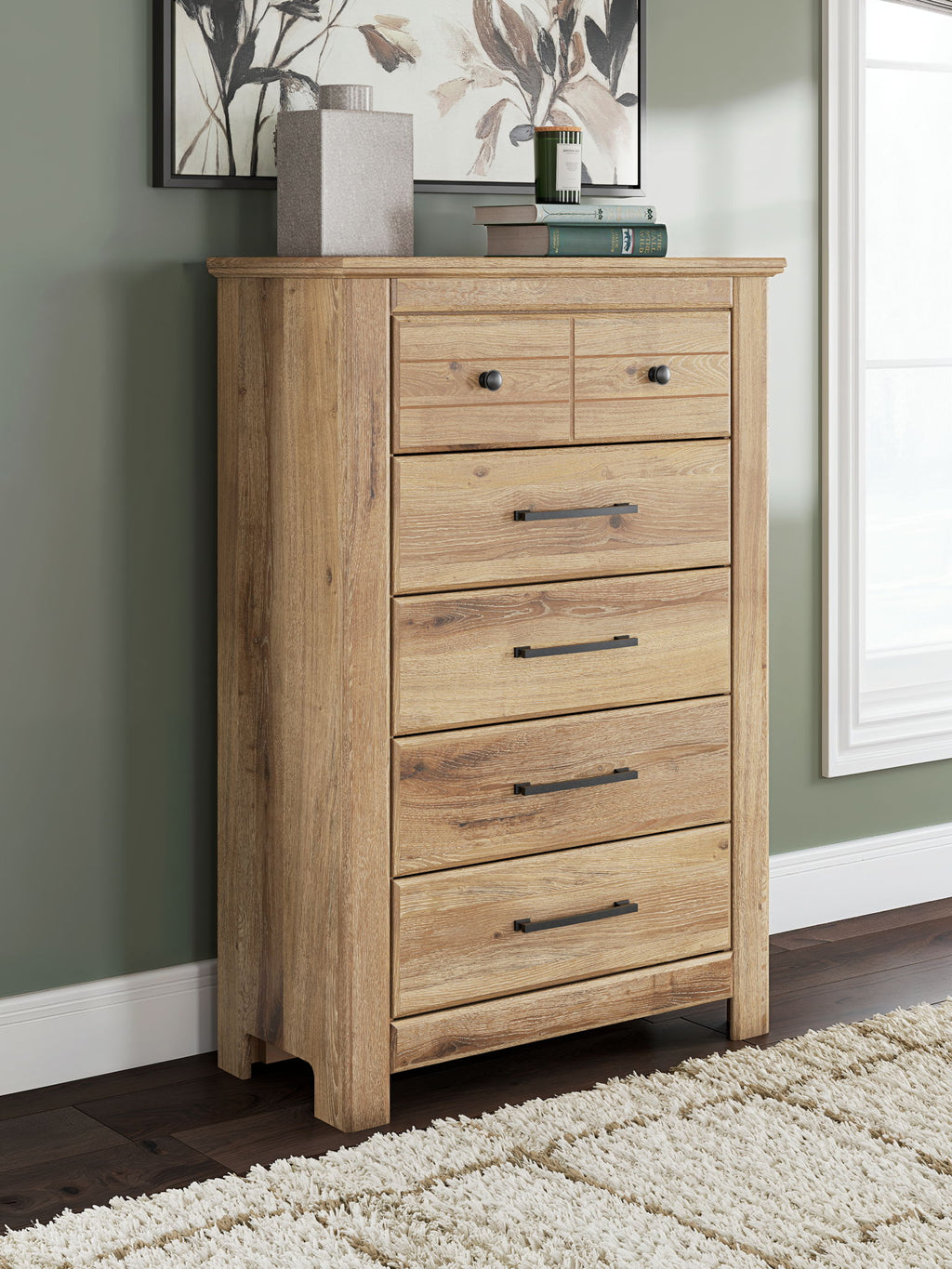 Light brown five-drawer chest with black bar pulls in a bedroom setting