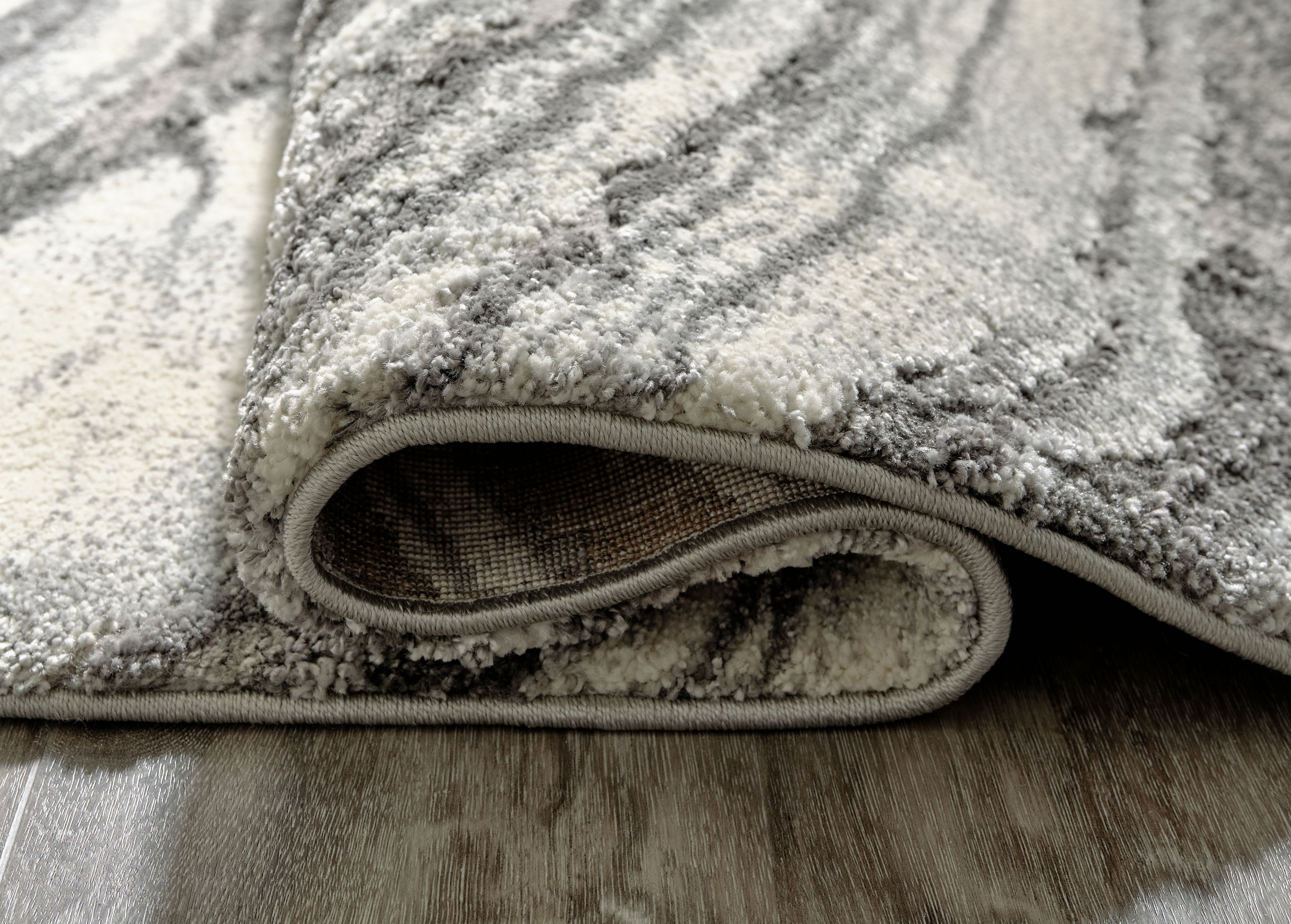 Rug corner close-up with folded edge and short textured pile in gray.