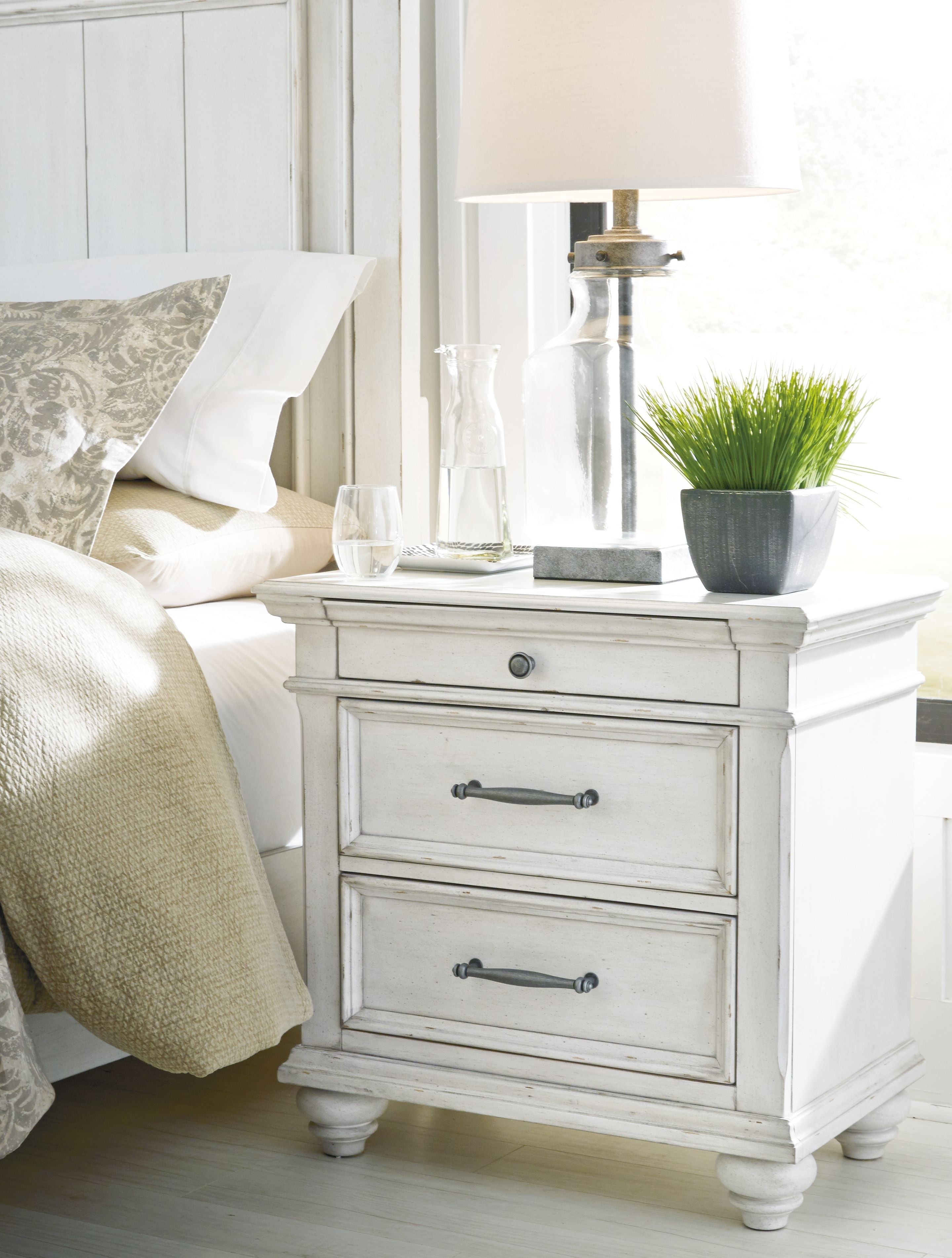 Whitewashed three-drawer nightstand with metal handles and turned legs, angled view