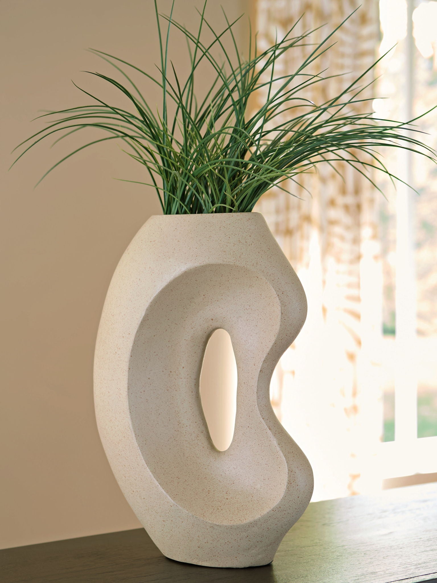 Beige ceramic vase with sculpted cutout, holding green grass stems.