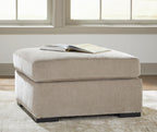 Oversized accent ottoman in light beige upholstered fabric with cushioned top and dark block legs.