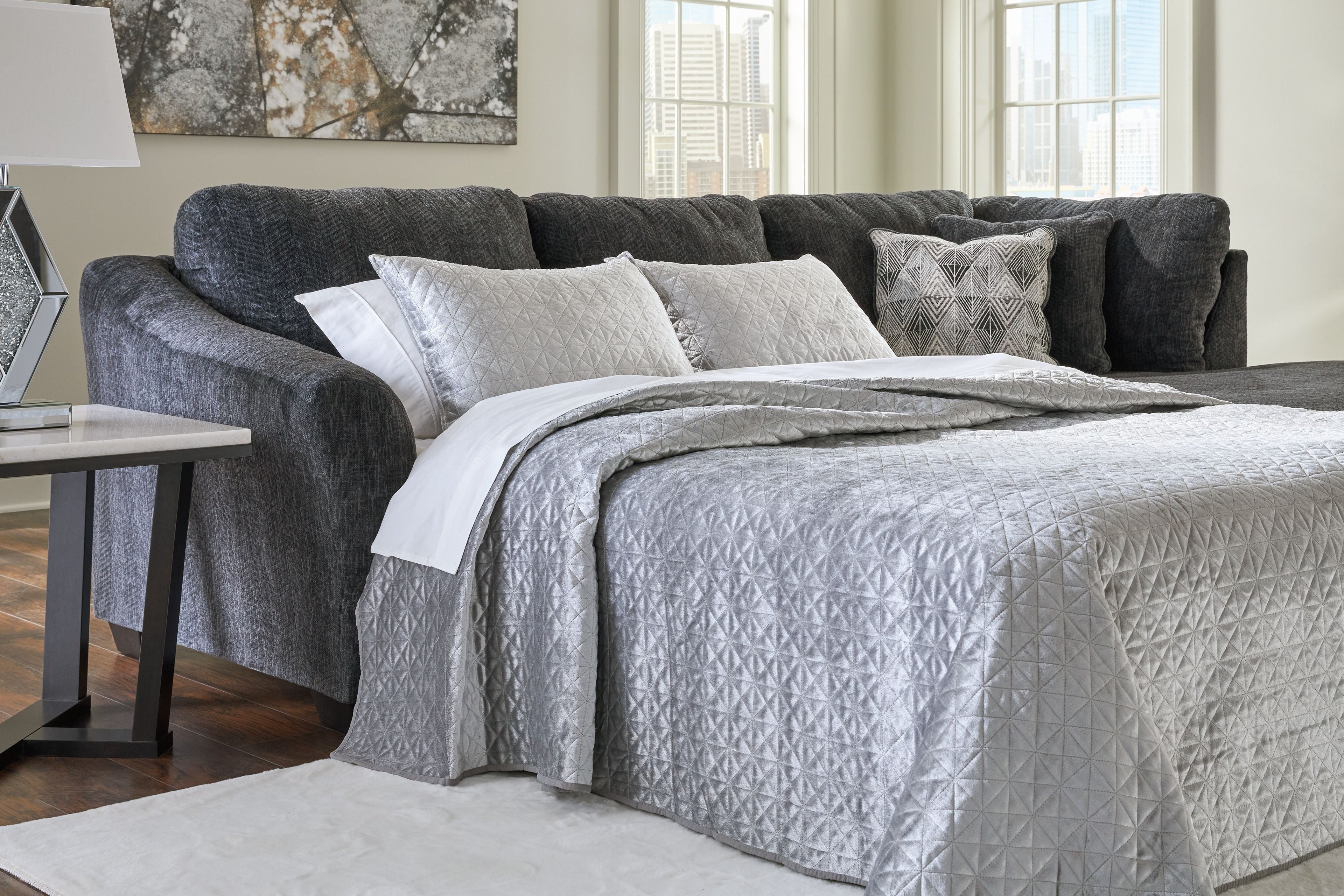 Dark gray sectional sleeper with open mattress, white quilt, and patterned throw pillows