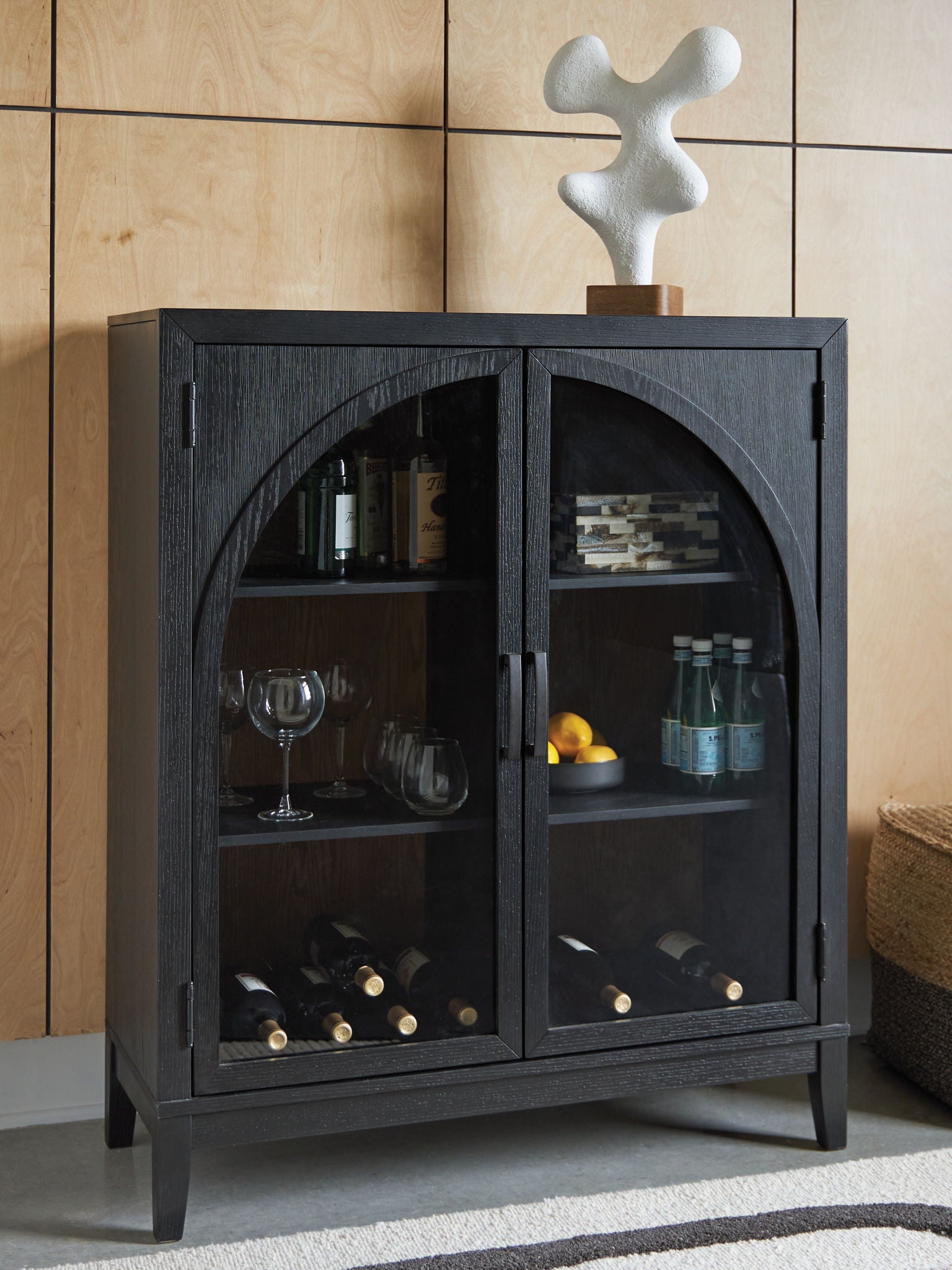 Black bar cabinet with arched glass doors showing bottles, stemware, and wine storage