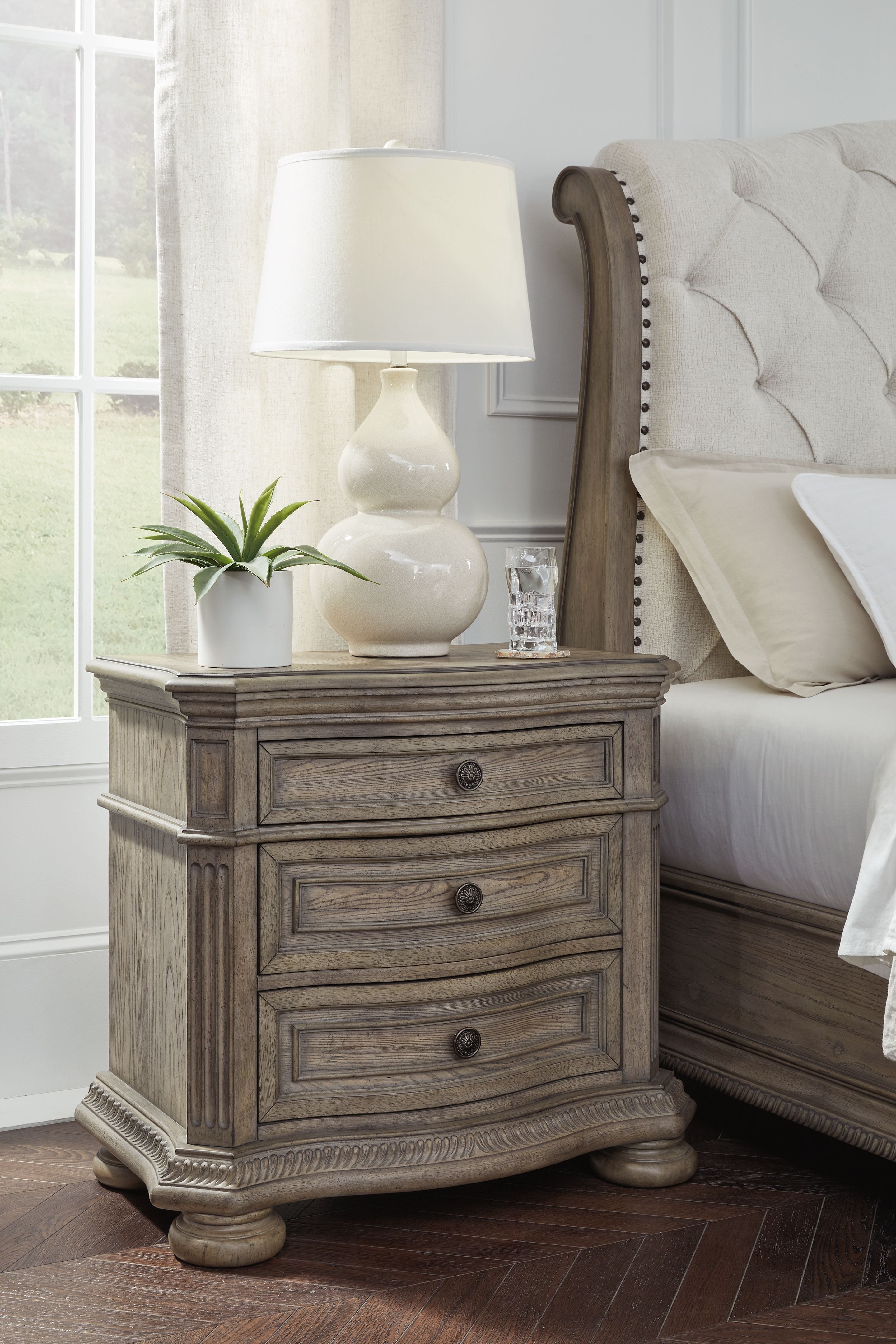 Light brown three-drawer nightstand with decorative carved base and turned legs