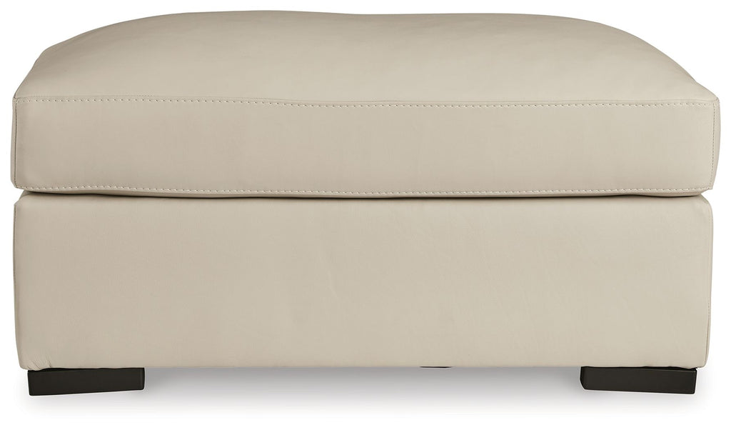 Almond padded ottoman with rounded top, visible panel stitching, and dark feet.