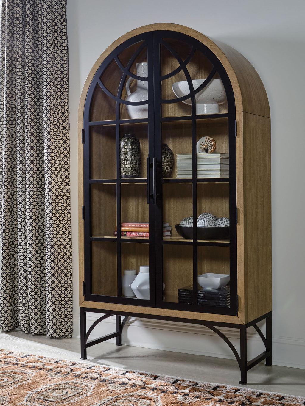 Natural wood accent cabinet with arched black grid glass door and items on shelves
