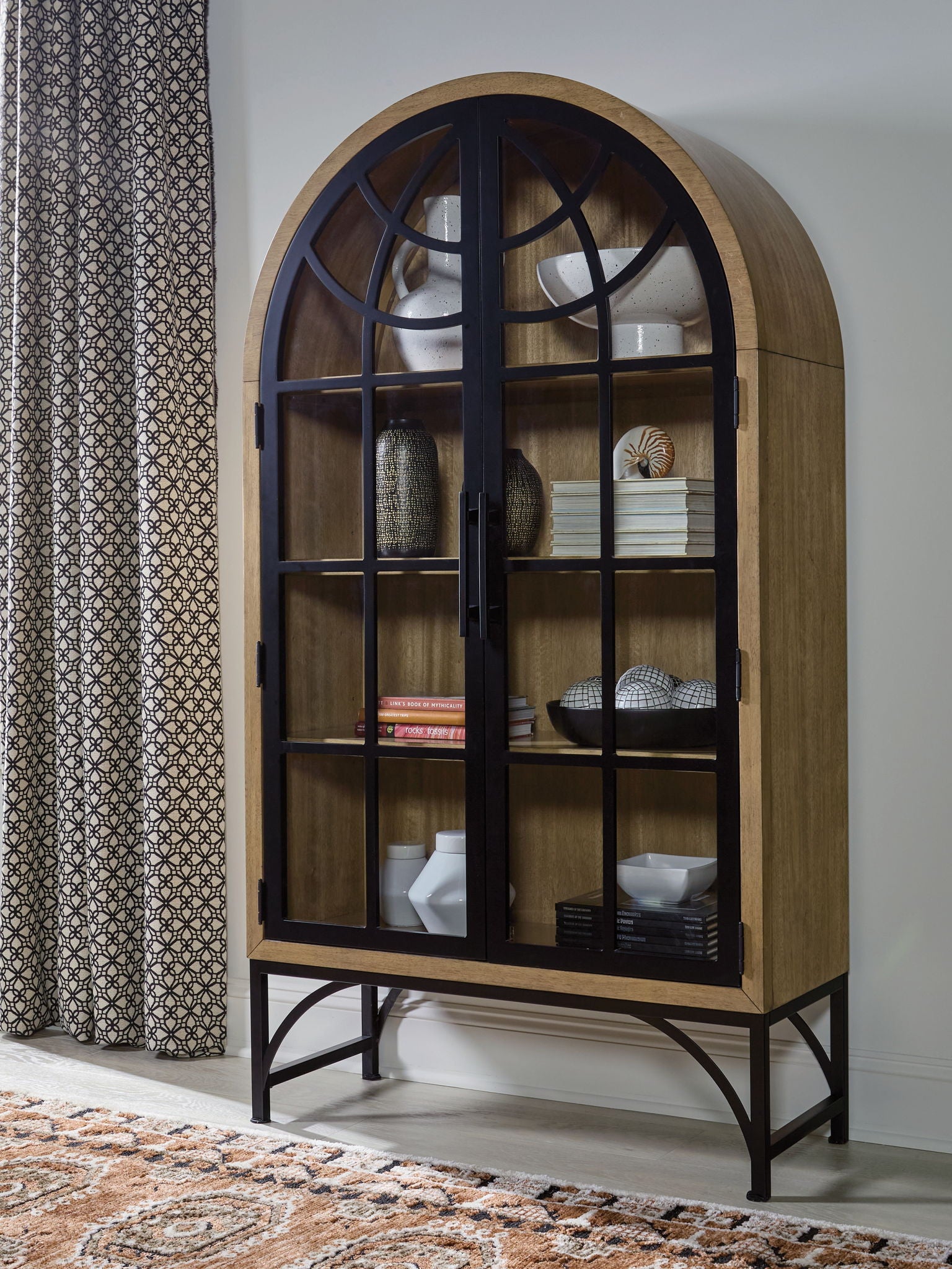 Natural wood accent cabinet with arched black grid glass door and items on shelves