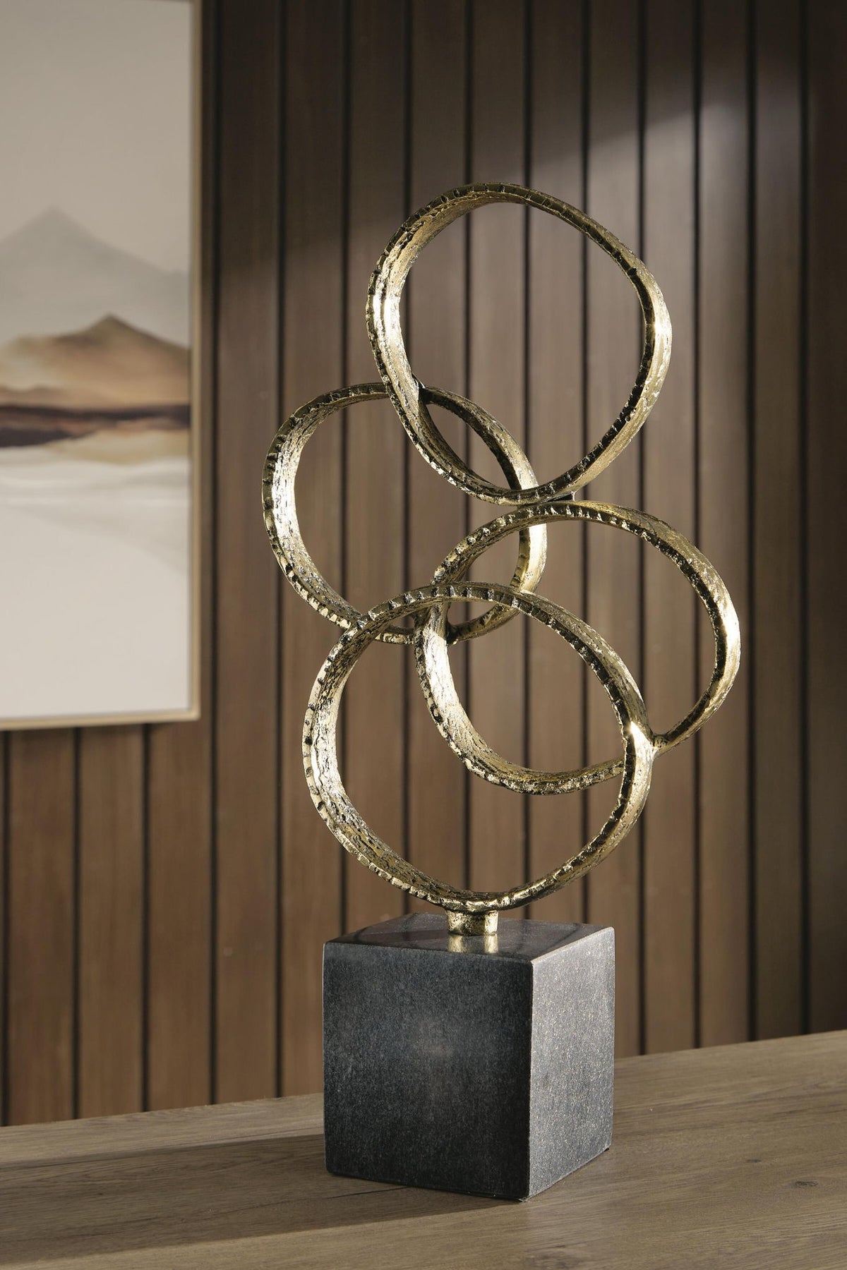 Gold-finish metal ring sculpture with three interlocking loops on dark pedestal