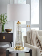 Glass table lamp with white drum shade, clear glass column, and gold base accents
