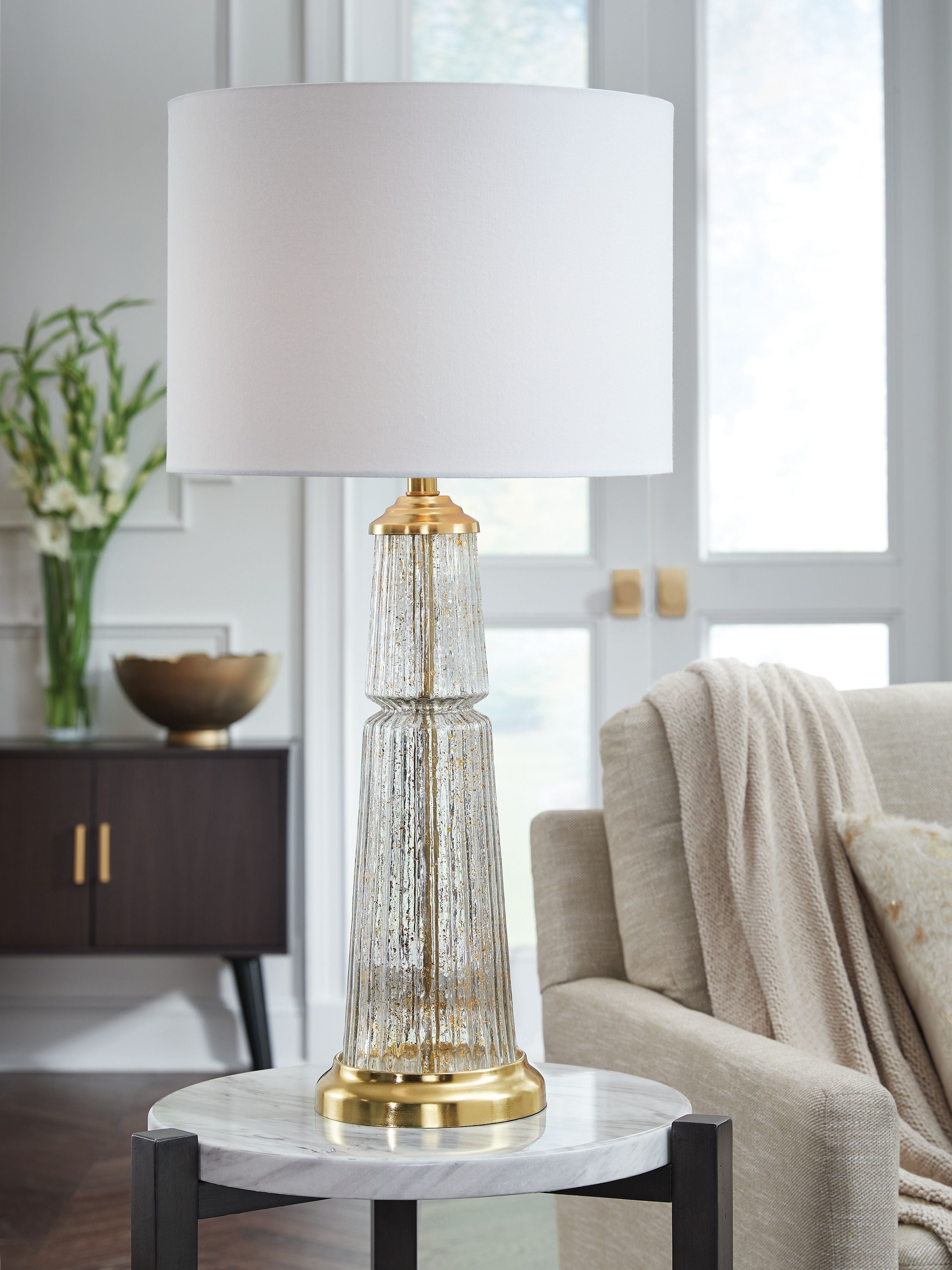 Glass table lamp with white drum shade, clear glass column, and gold base accents