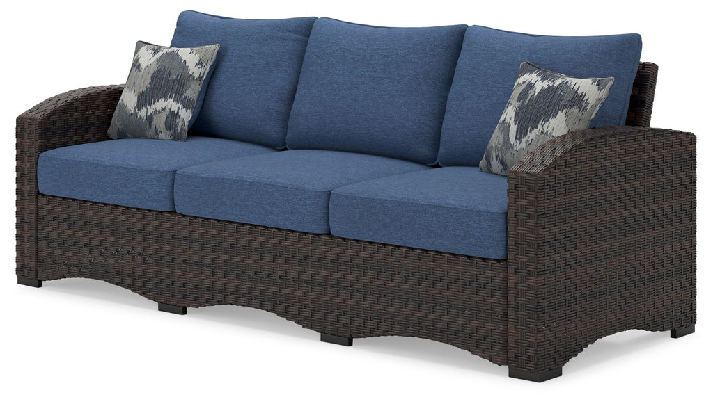 Outdoor sofa with blue cushions, brown wicker arms, two patterned throw pillows
