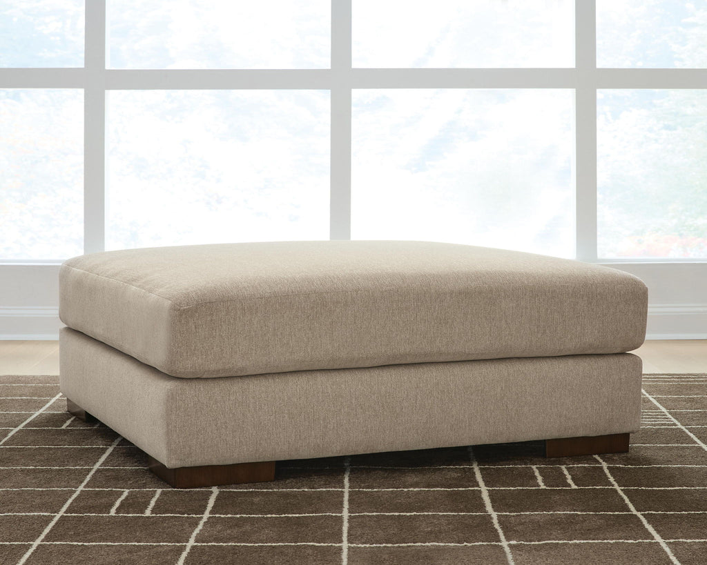 Oversized accent ottoman in dune beige with rounded lid, upholstered sides, and dark feet.