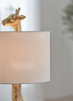 Gold table lamp close-up with giraffe head and cream fabric drum shade.