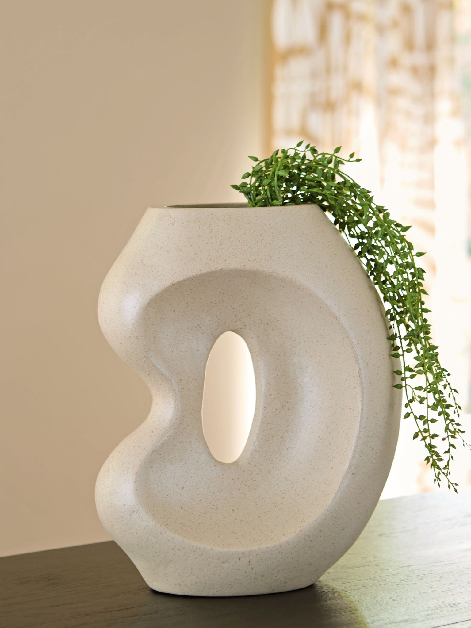 Beige ceramic vase with oval cutout, topped with trailing green plants.