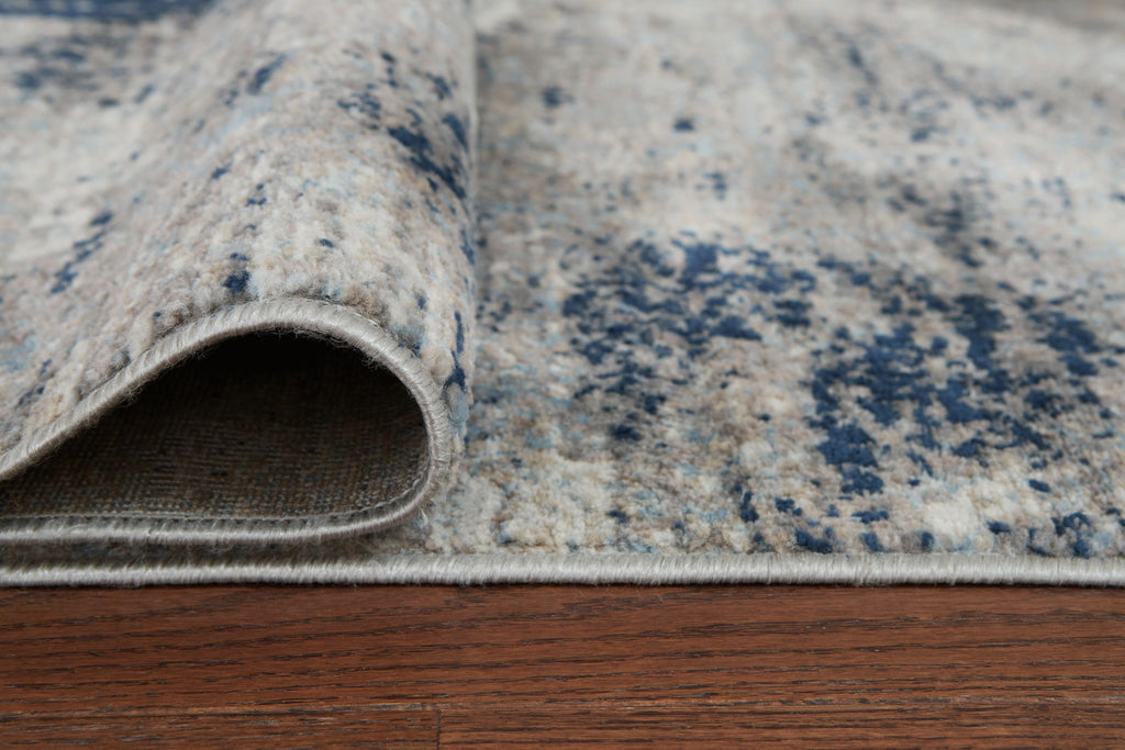 Rug corner view with rolled edge and gray pile covered in blue speckled pattern