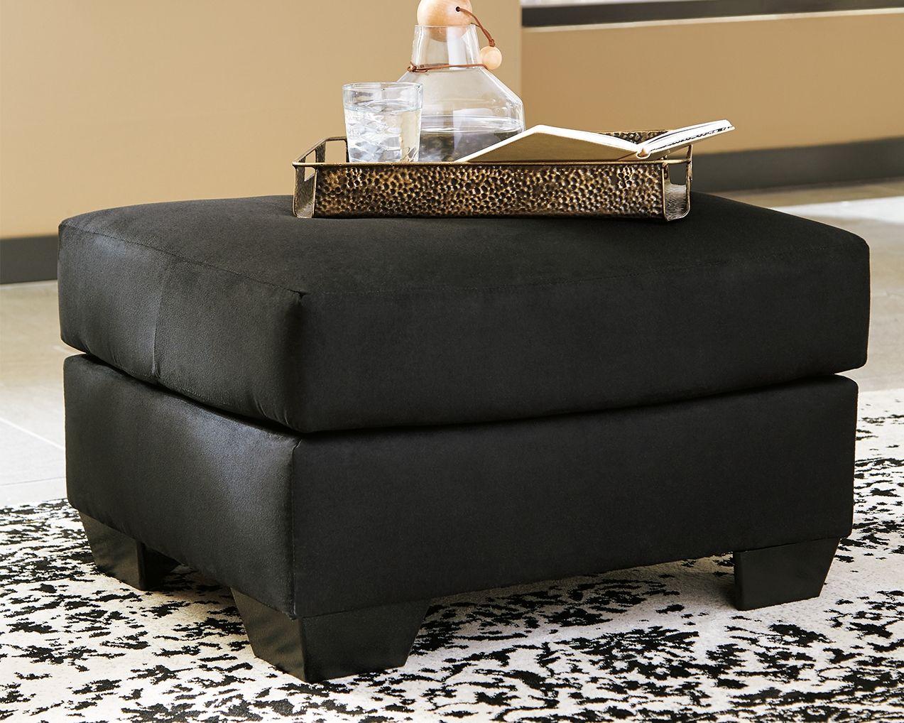 Upholstered ottoman in black fabric, thick padded top with short legs.