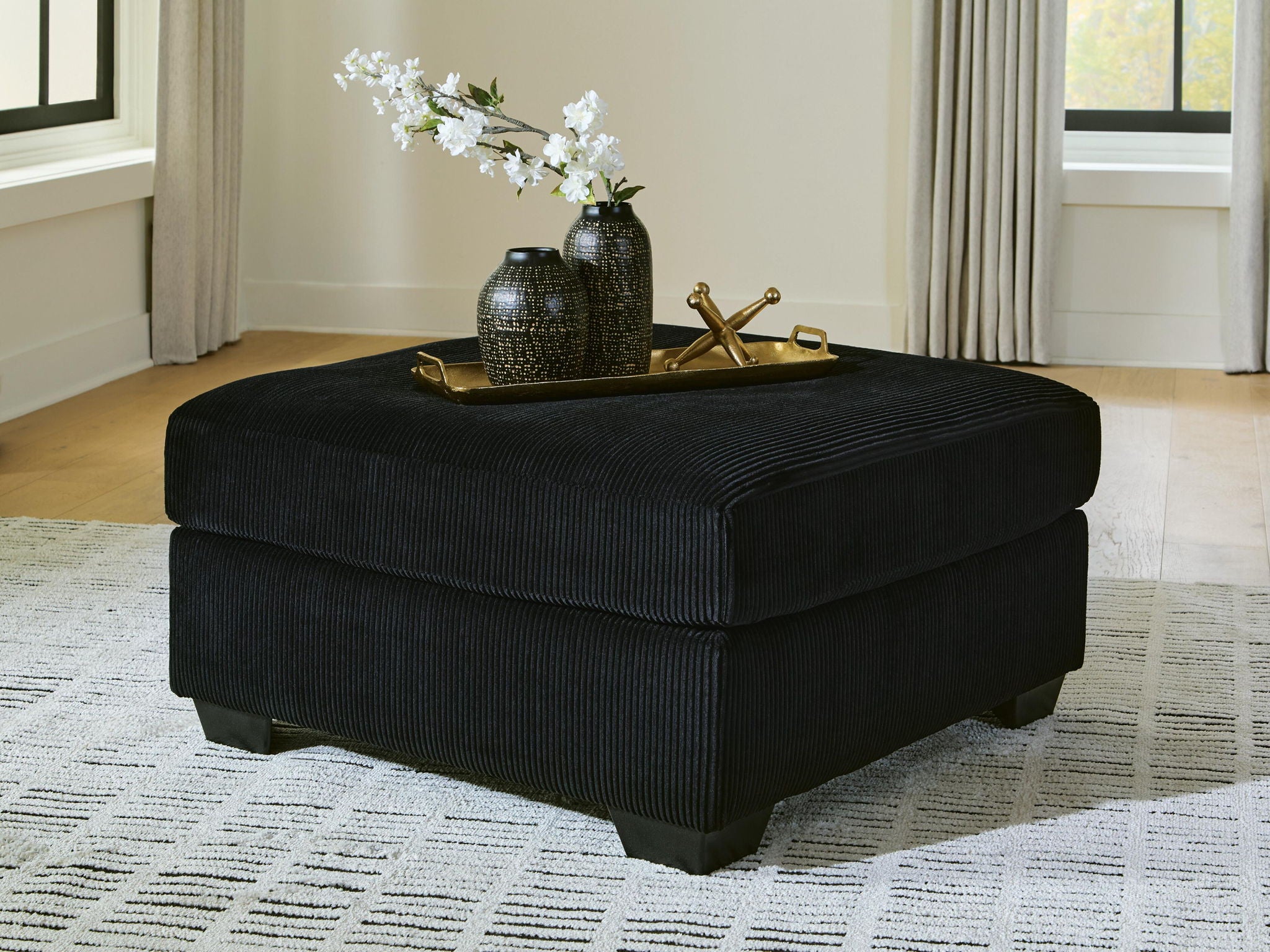 Black oversized ottoman with ribbed fabric, square padded top, dark legs.