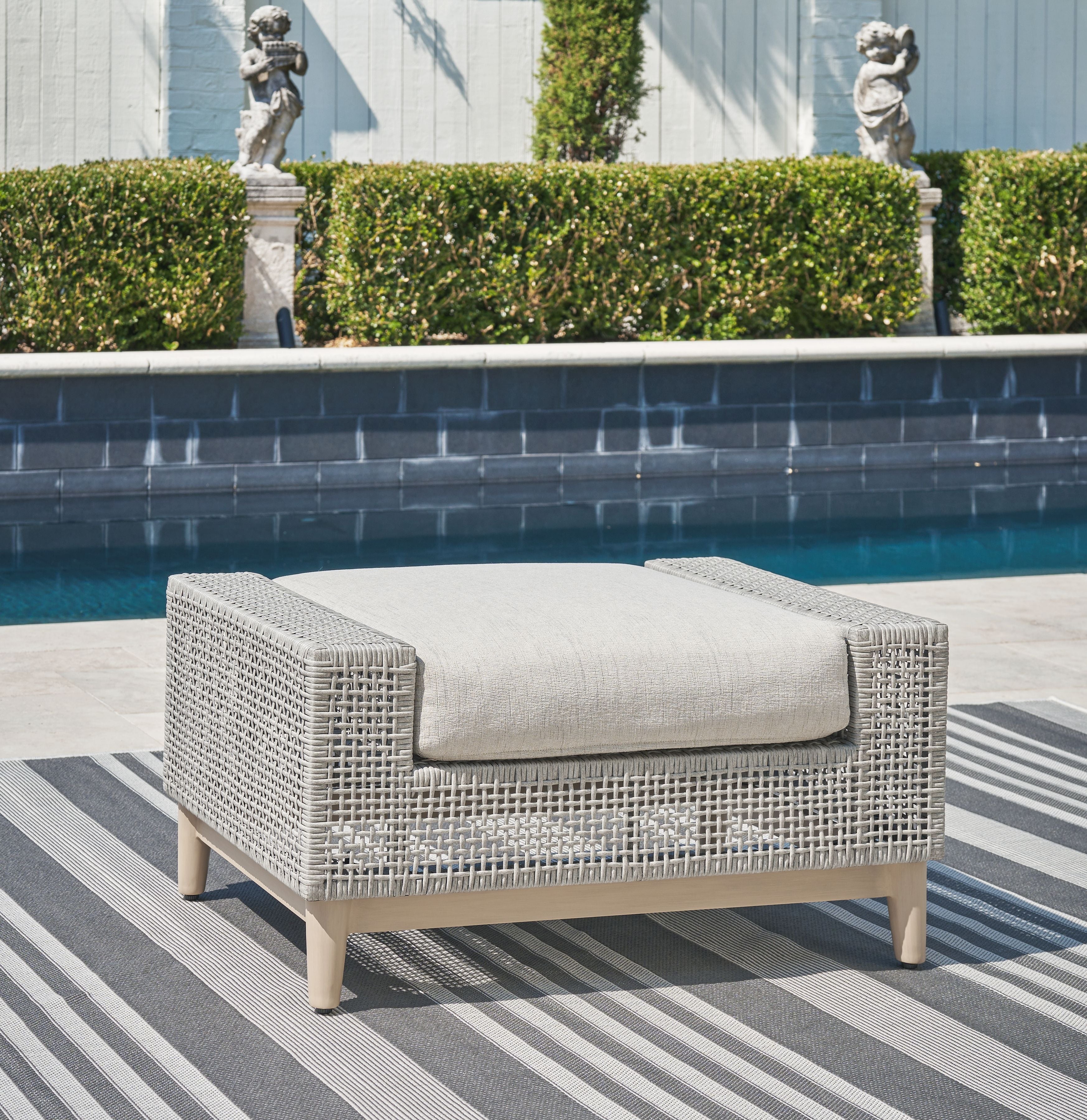 Gray outdoor ottoman with woven sides and padded cushion on light wood legs