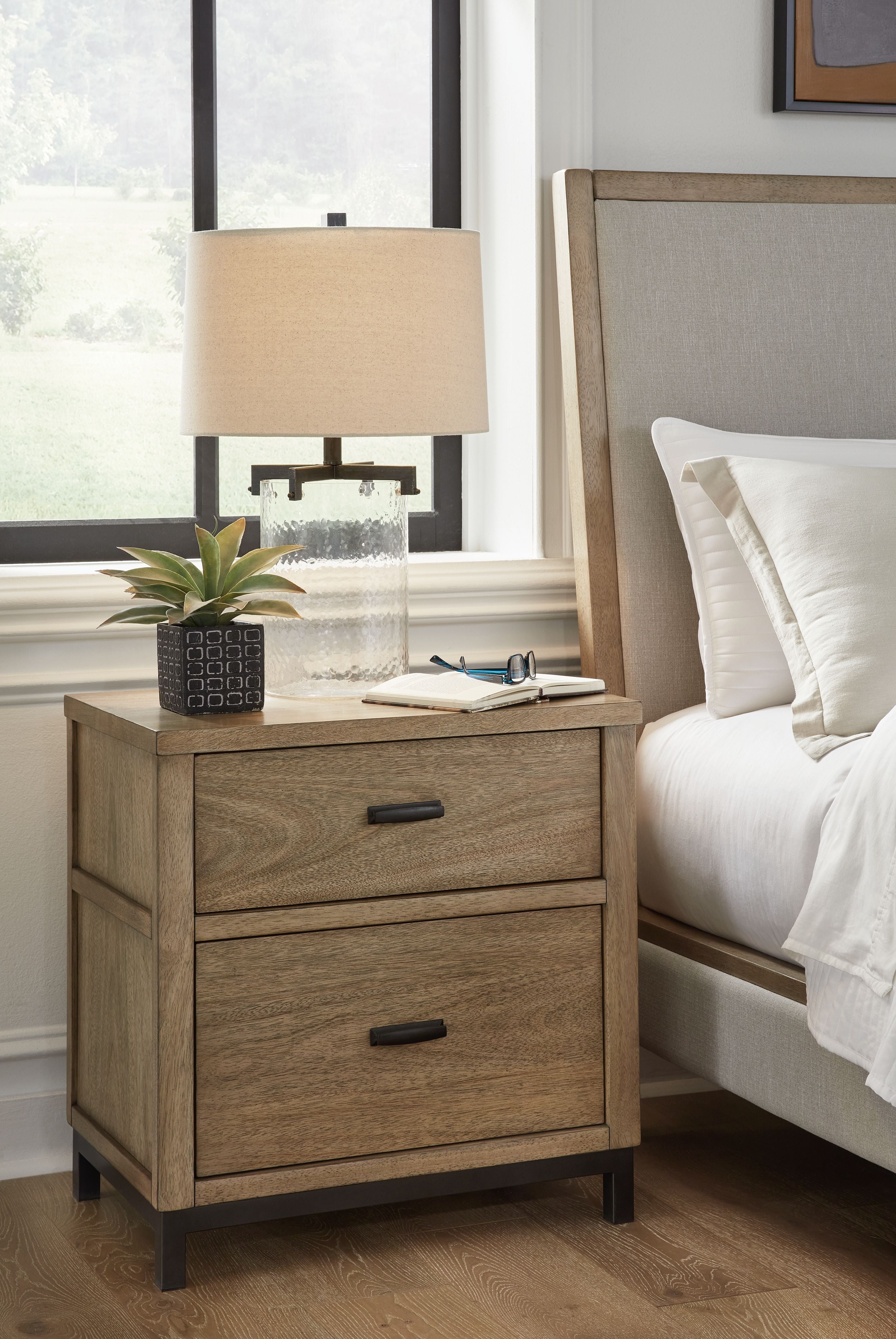 Light brown two-drawer nightstand with black handles shown next to a bed