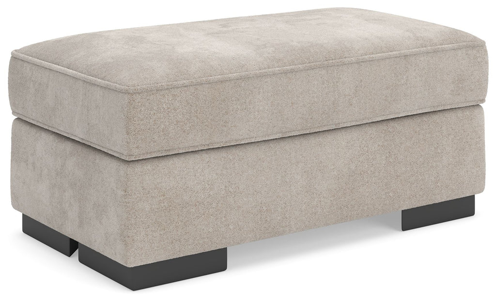 Taupe upholstered ottoman with padded top and black base legs, rectangular profile