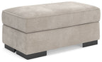 Taupe upholstered ottoman with padded top and black base legs, rectangular profile