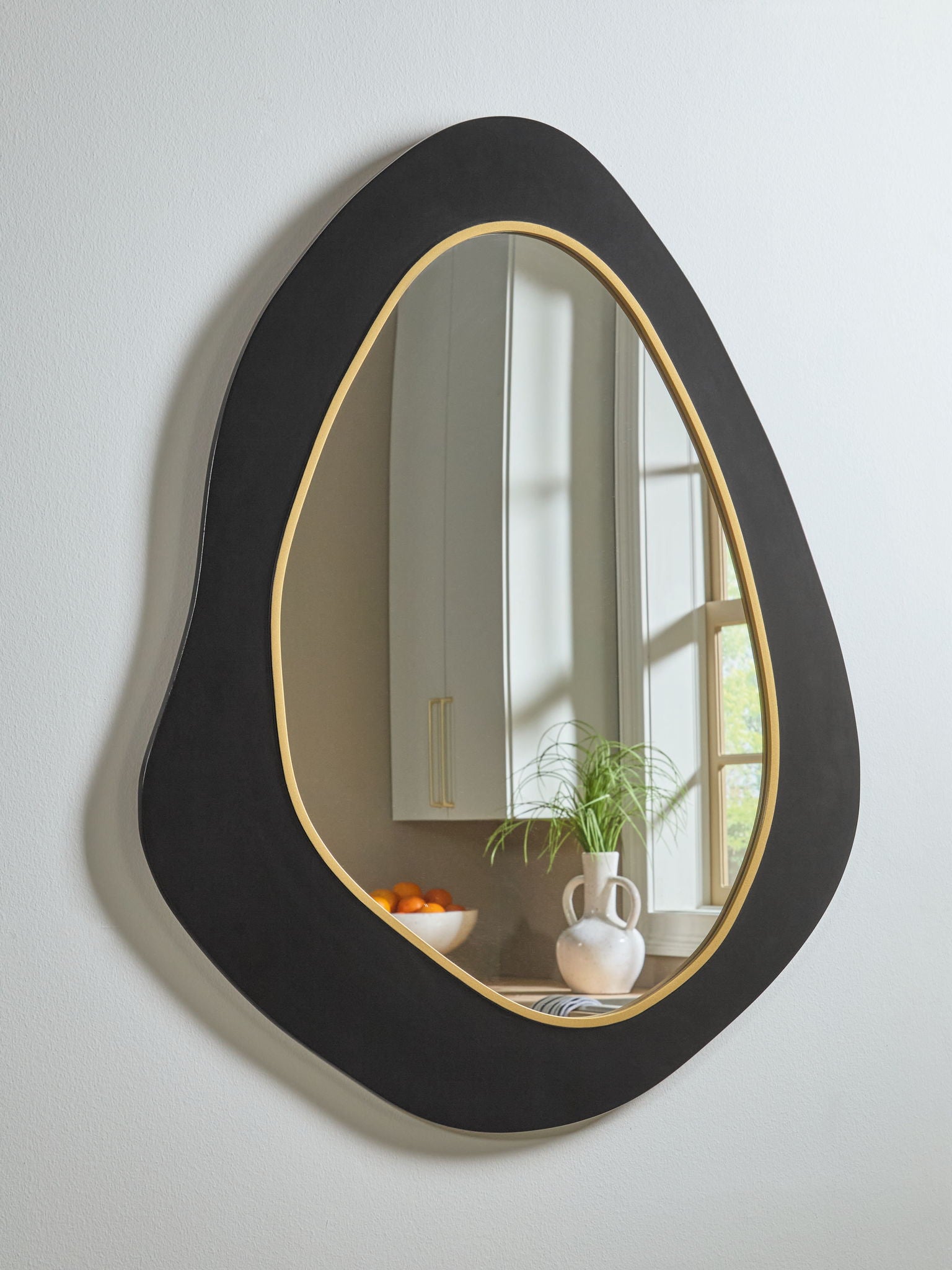 Black oval wall mirror with gold rim, reflecting window light and decor