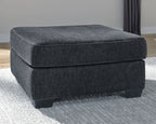 Dark gray upholstered accent ottoman with padded seat, rounded edges, and short legs